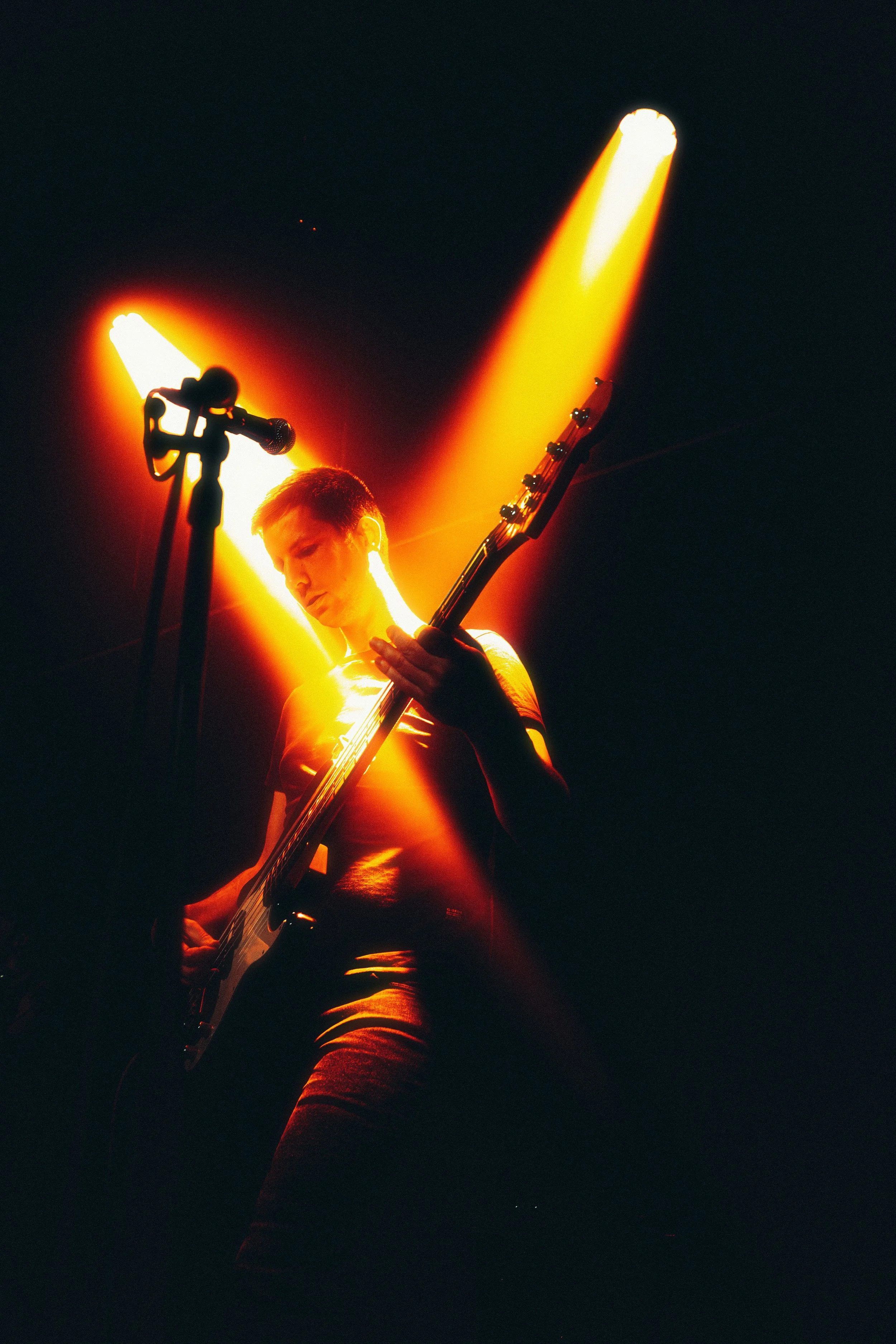 A young person playing an electric guitar on stage, illuminated by bright yellow stage lights against a dark background.