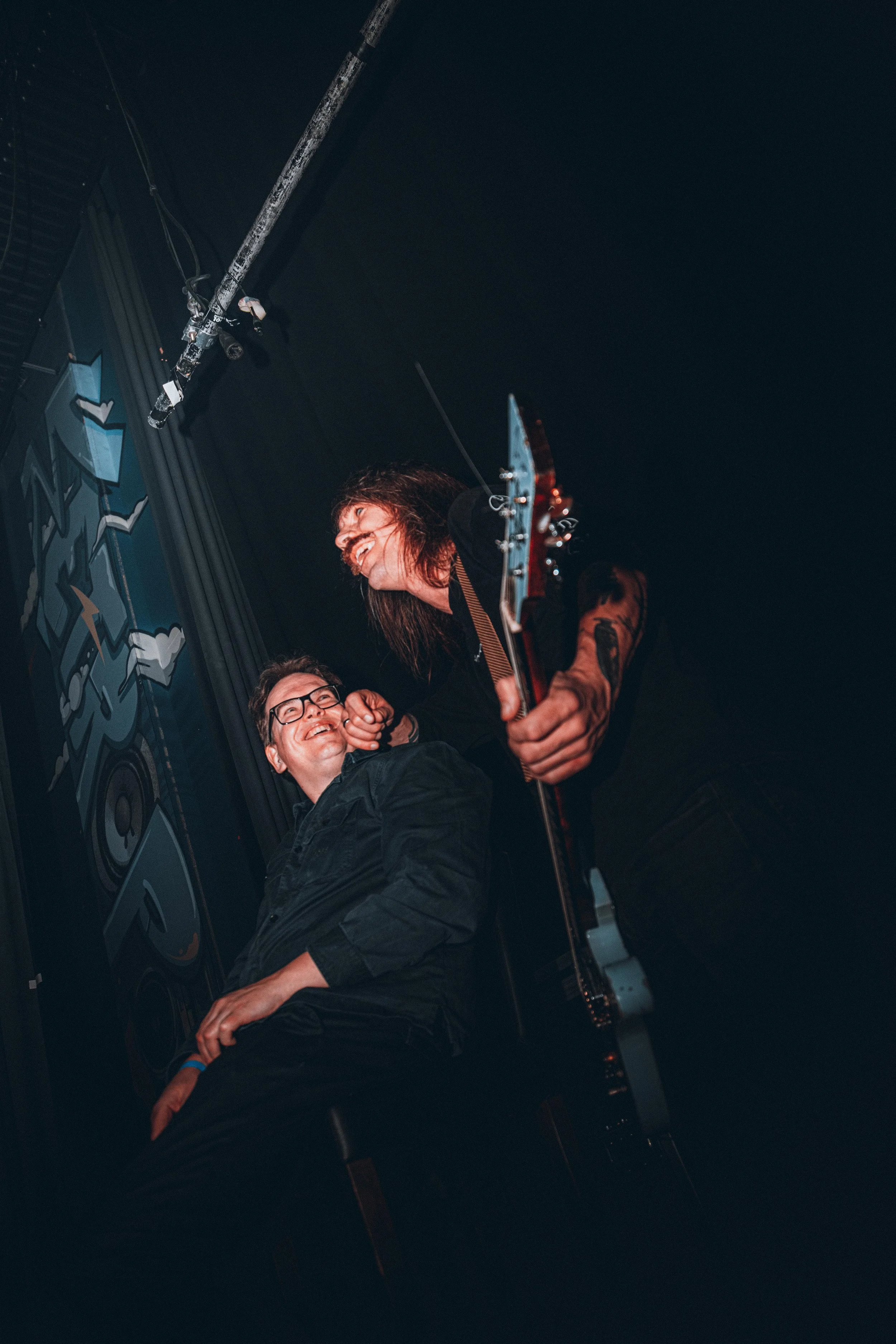 Two friends share a joyful moment during a live music performance, one is playing an electric guitar while the other is sitting and smiling, with a stage backdrop behind them.