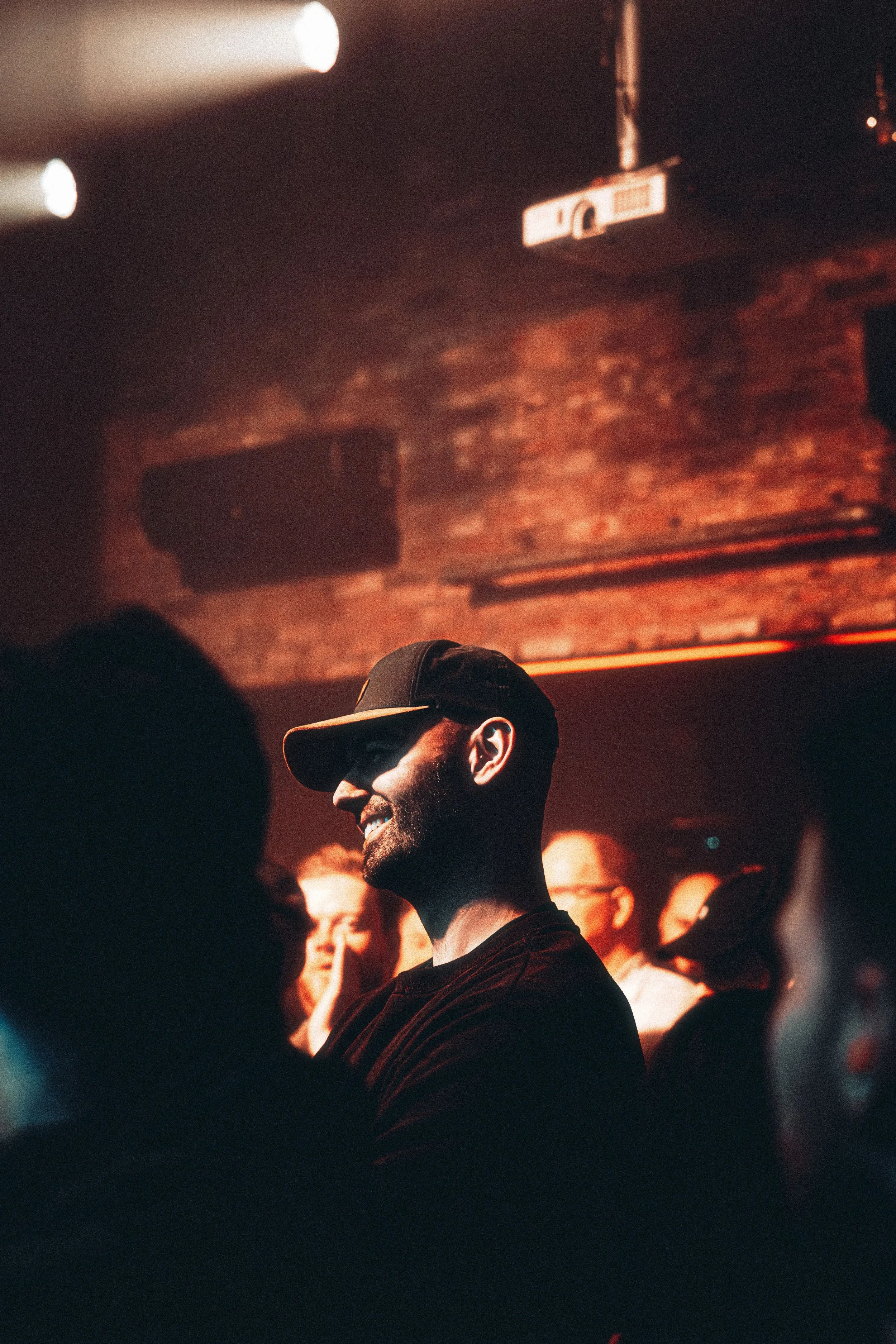 A man wearing a cap and smiling at a crowded indoor event with exposed brick walls and warm lighting.