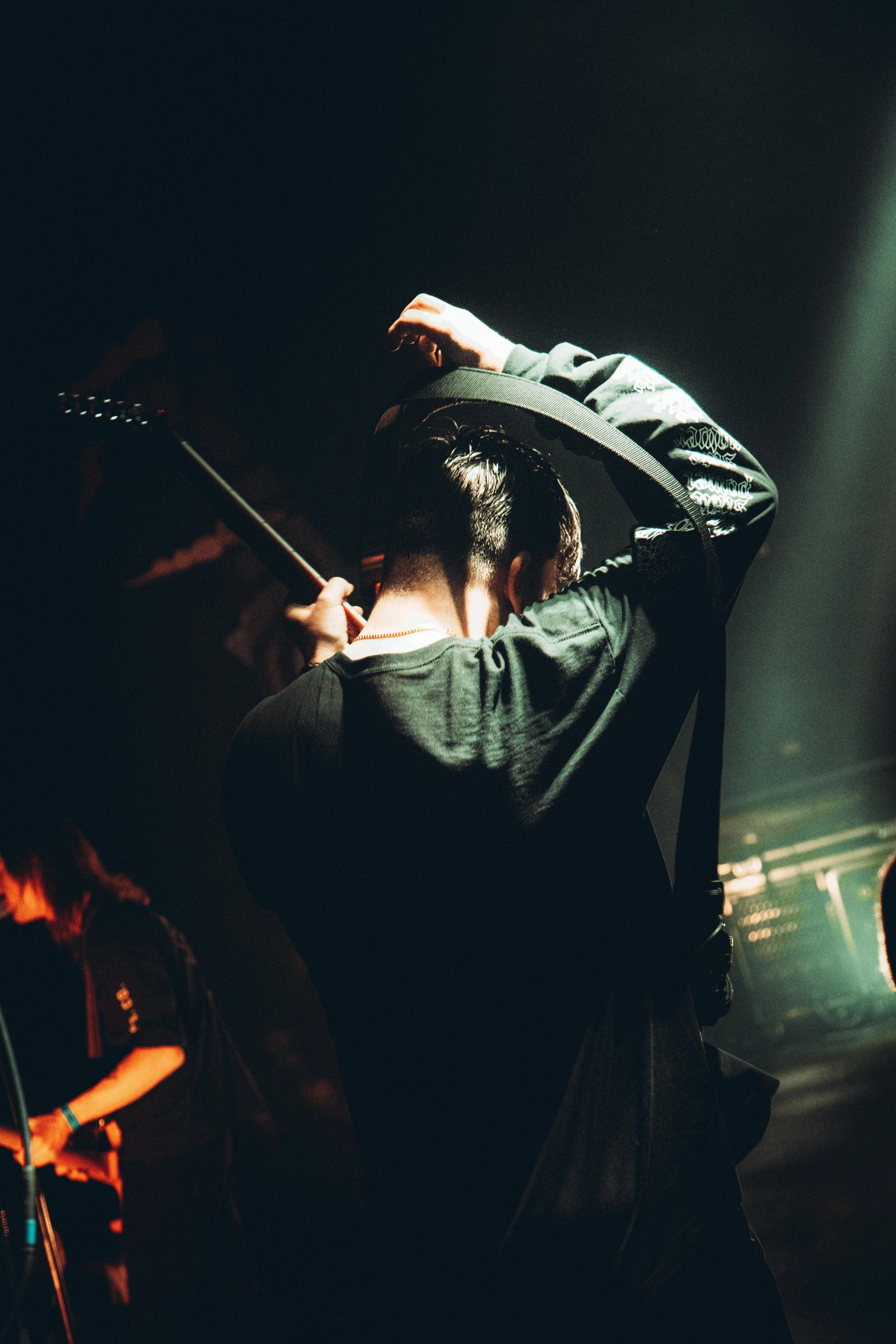 A person adjusting a guitar strap on their shoulder backstage or in a dimly lit area.