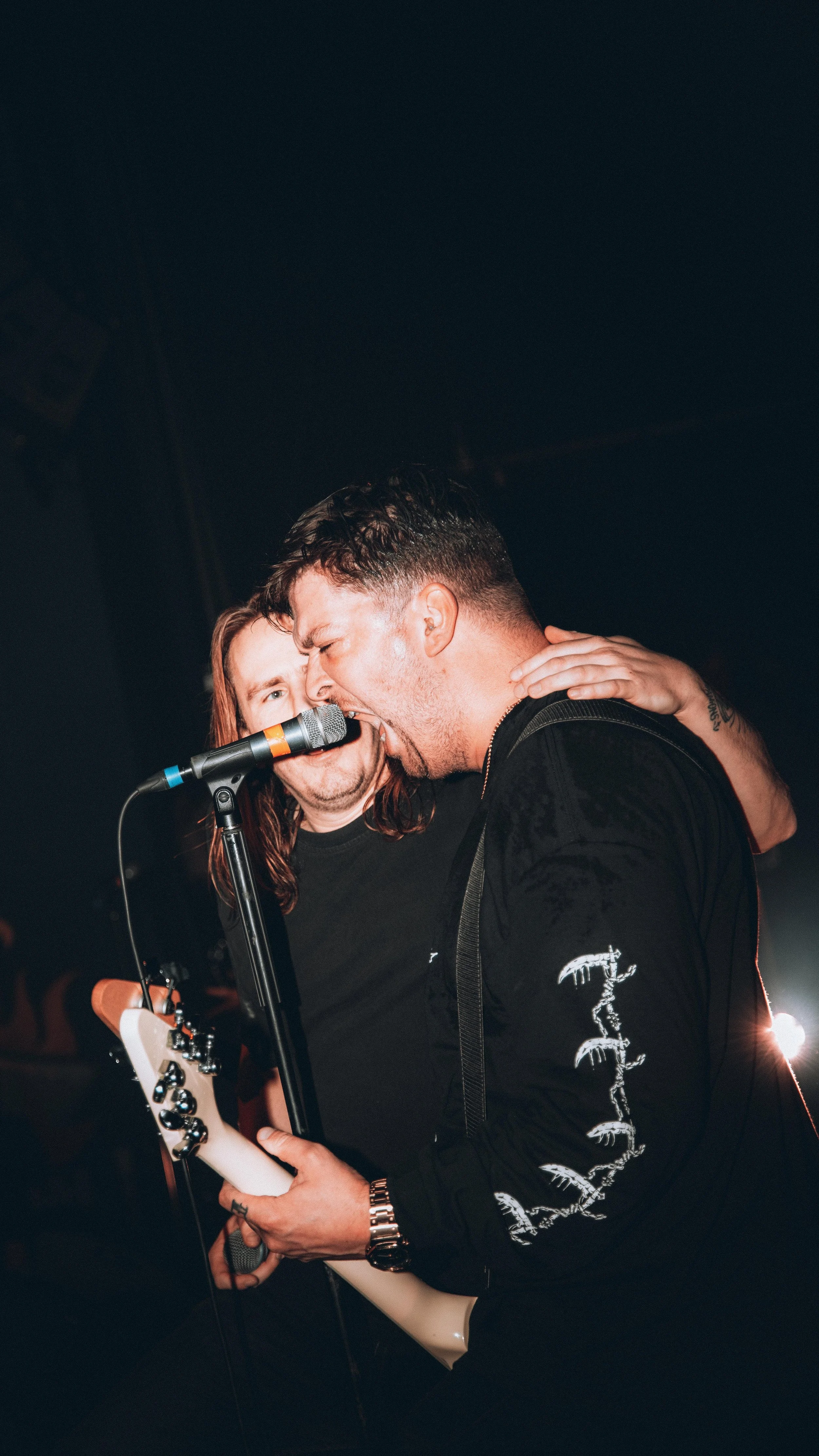 Two male musicians singing into a microphone during a live performance, one playing an electric guitar and the other supporting with their hand on the guitarist's shoulder.