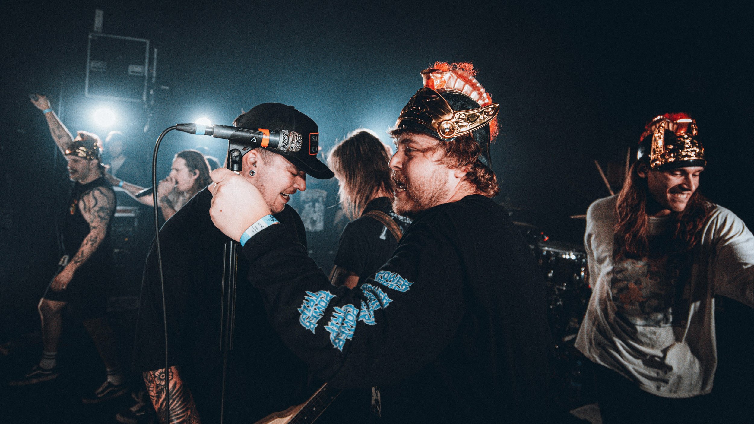 Group of people on stage at a concert, wearing costumes and accessories like helmets and hats, dancing and enjoying music with stage lights in the background.