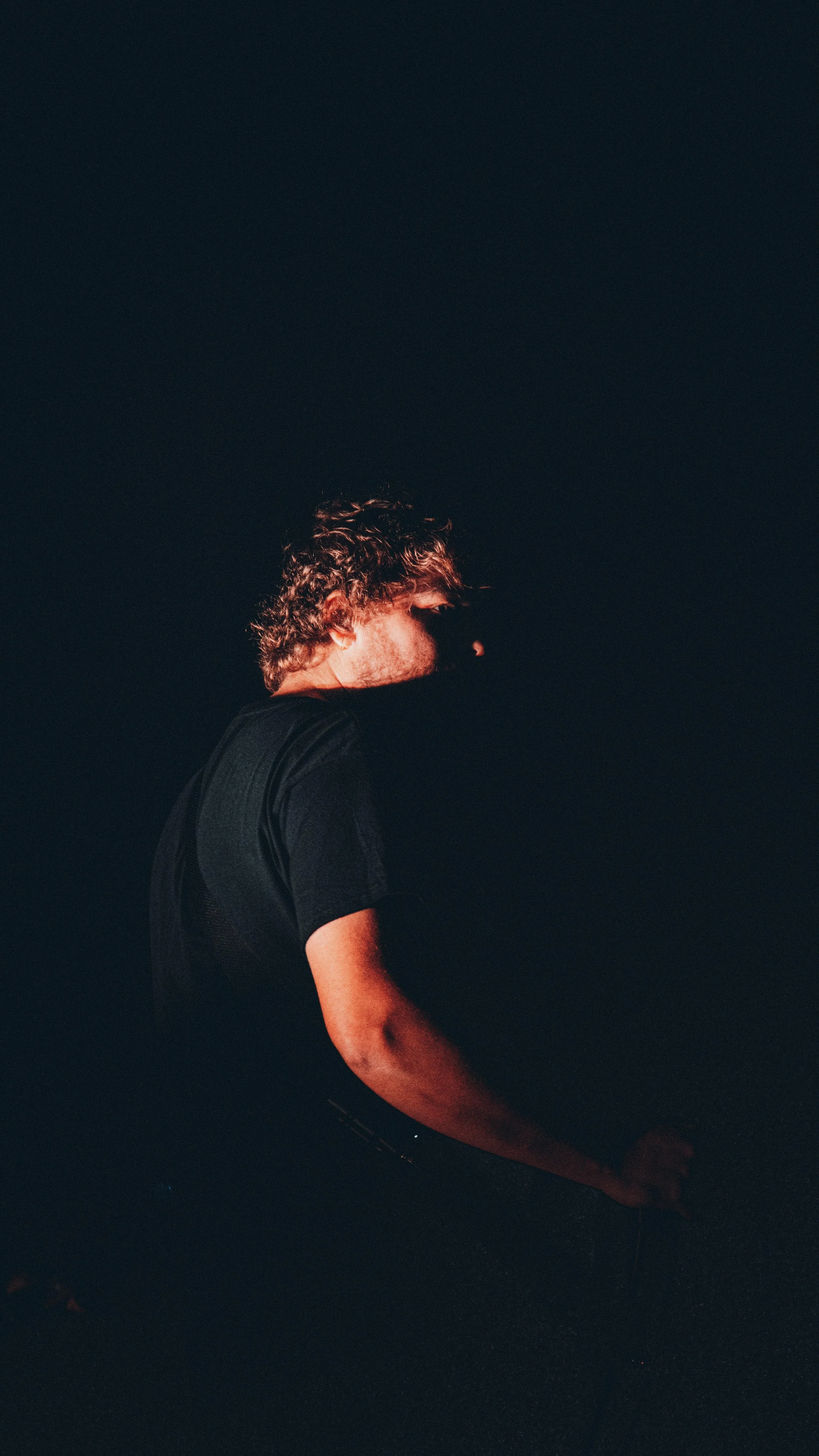 A person with curly hair wearing a black shirt, mostly obscured by darkness, with only part of their body and face visible in low lighting.