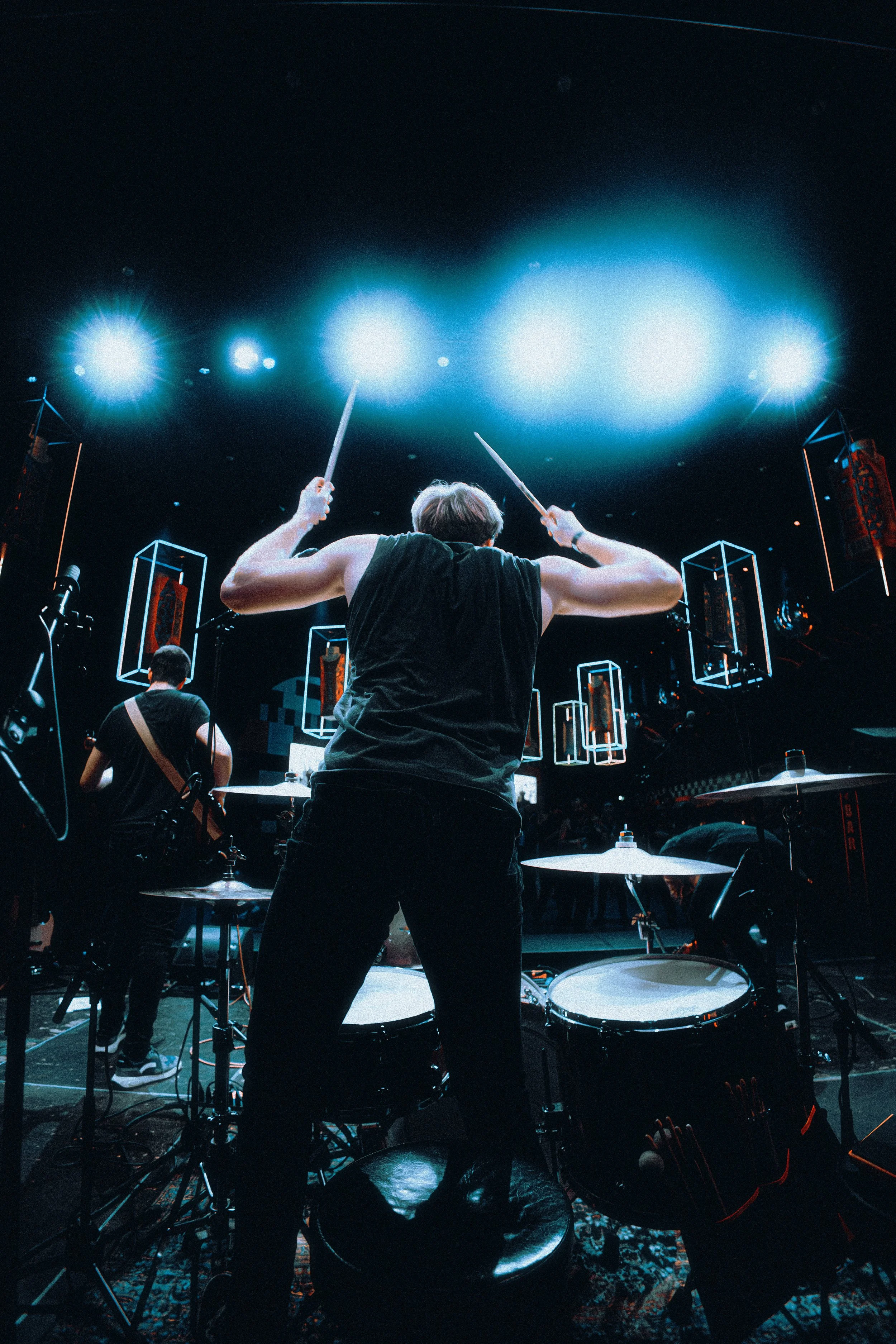 A drummer playing on stage with bright lights overhead, and other band members visible in the background.