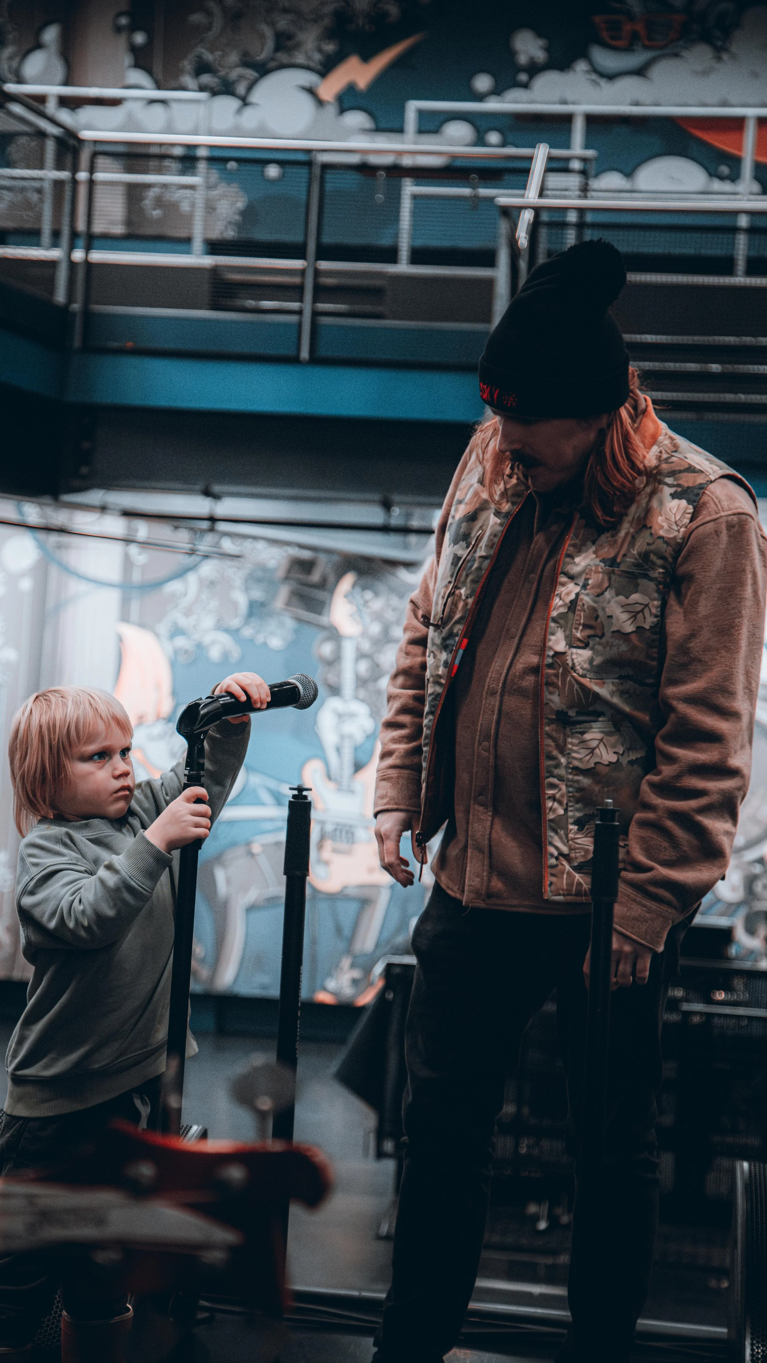 A young boy with blonde hair holding a microphone directed towards a man in a cap at an indoor music venue.