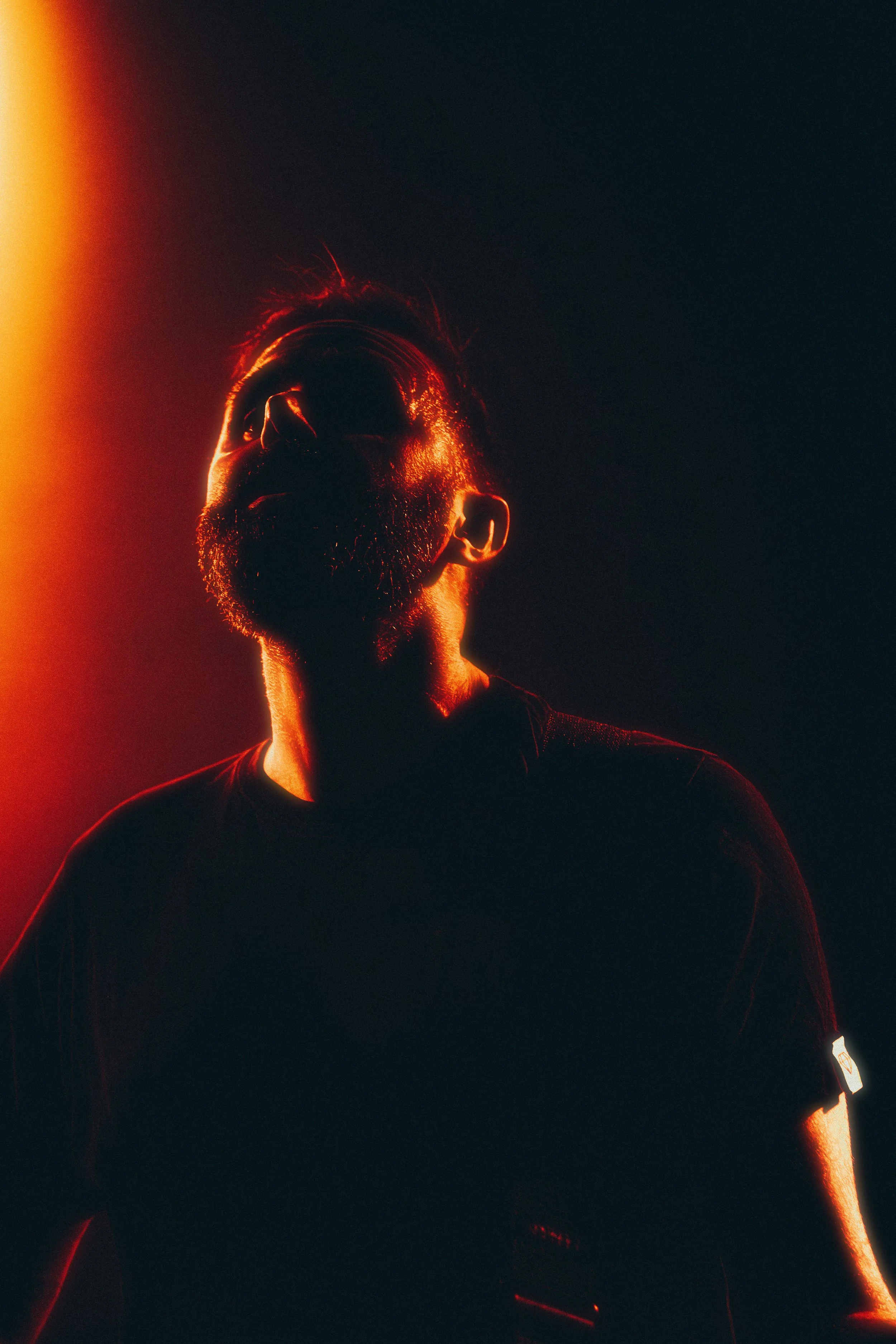 A man with a beard and hair styled up, illuminated by a red-orange light from the left side, against a dark background.