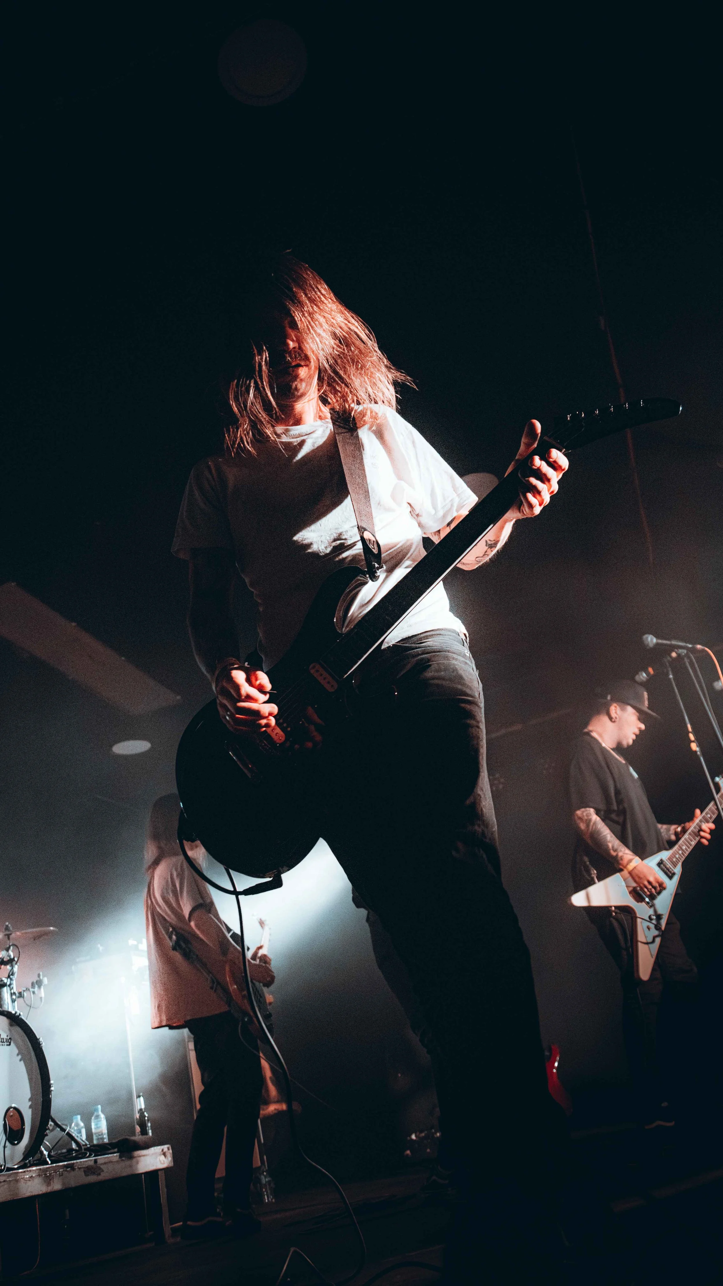Musicians performing live on stage, with the lead guitarist in the foreground playing an electric guitar, hair covering part of their face, and stage lights creating a dramatic atmosphere.