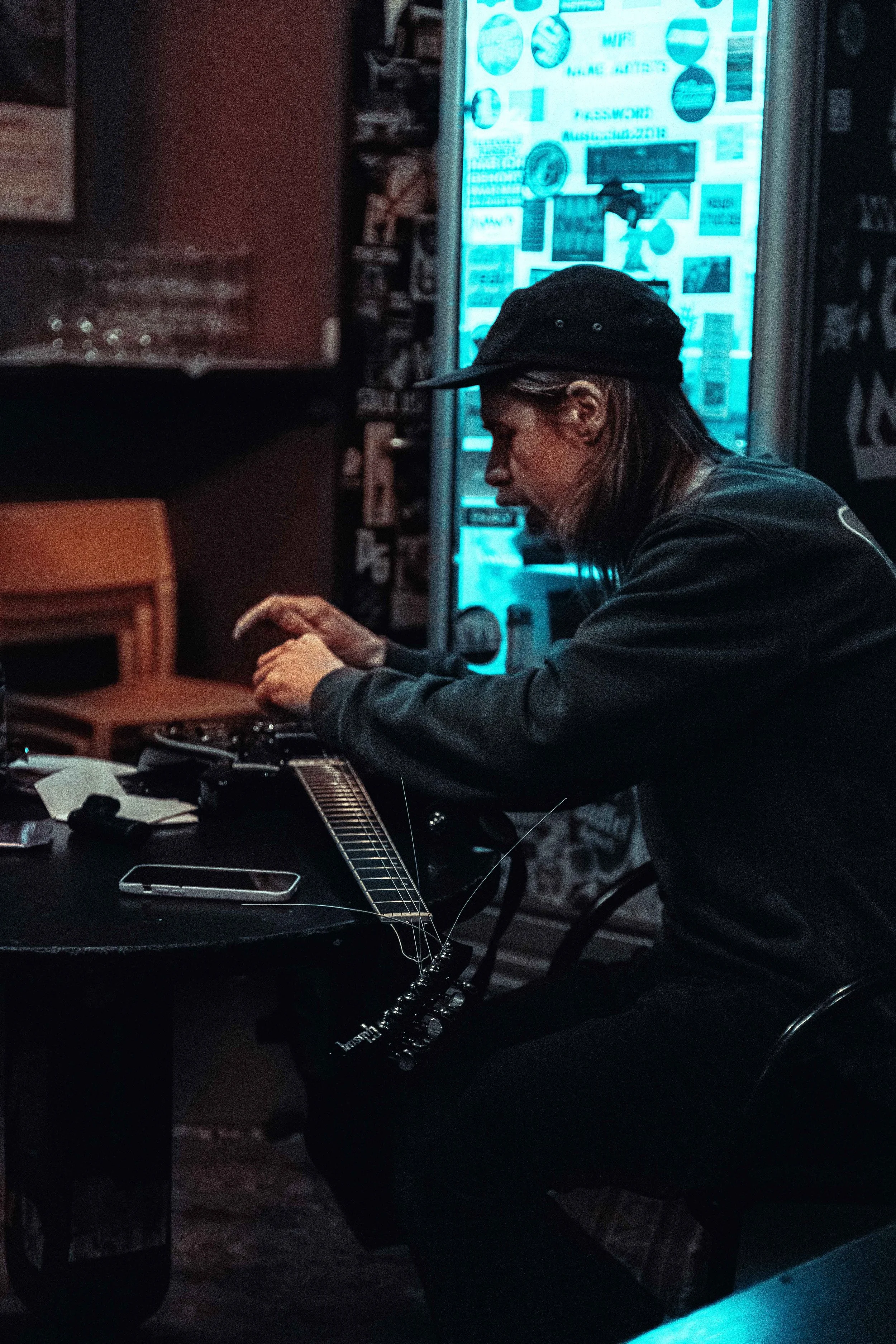 A man with long hair, wearing a black cap and black jacket, sitting at a table playing an acoustic guitar with a smartphone next to him in a dimly lit room with a blue-lit vending machine in the background.