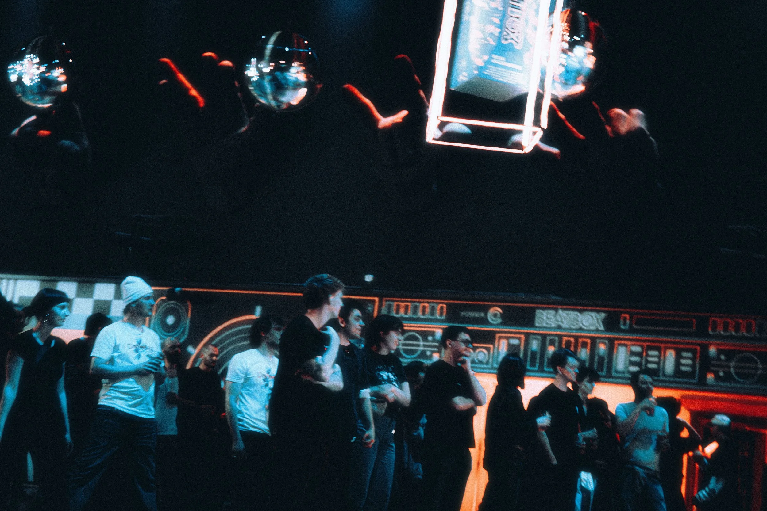 People at a futuristic exhibition or event, standing in front of a wall with neon lights and digital displays, and a large digital screen hanging above showcasing reflections of the scene.