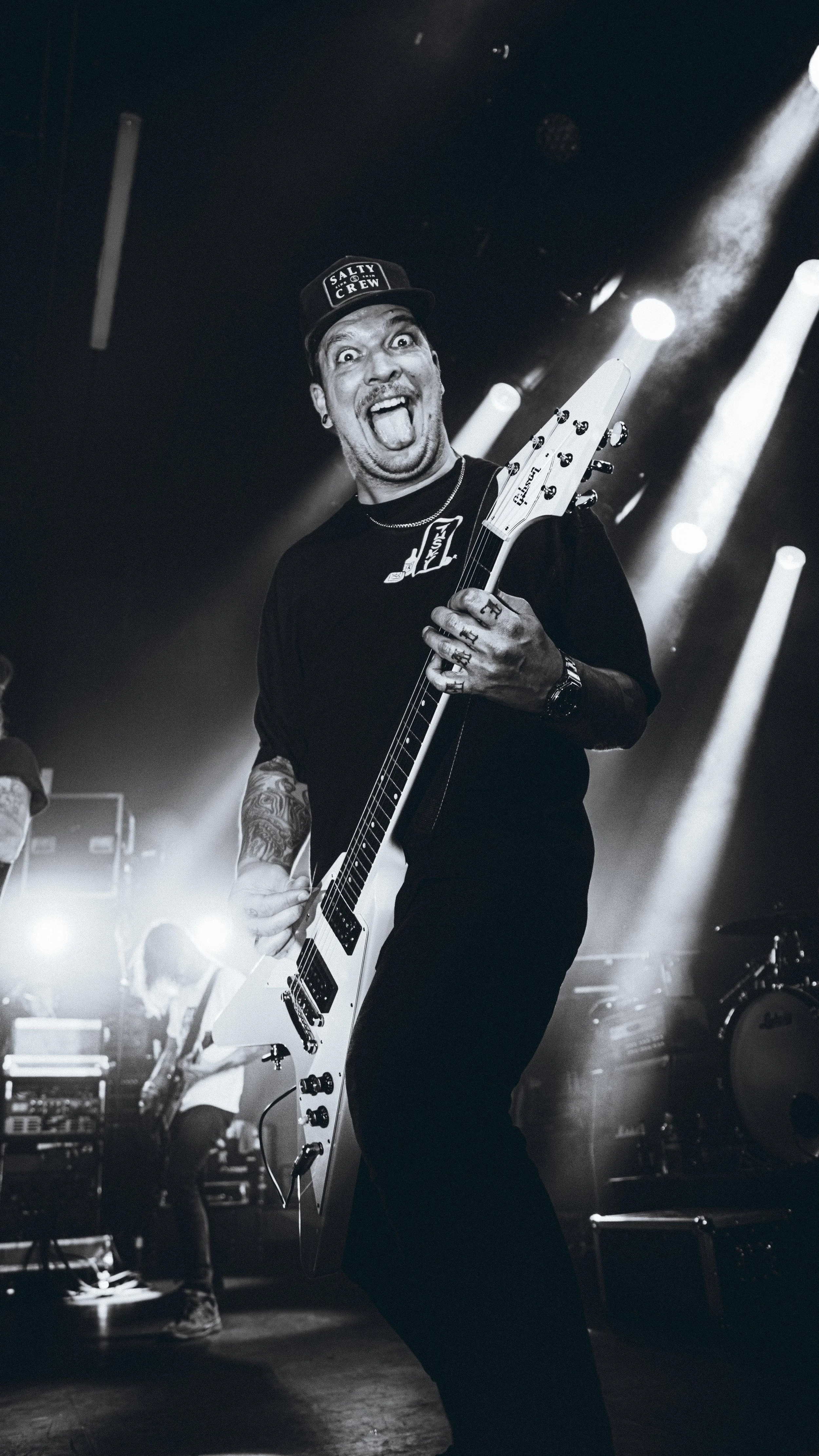 Black and white photo of a lively musician playing an electric guitar on stage, smiling with tongue out, wearing a black t-shirt and a hat that says 'Salty Crew', with stage lights and another band member in the background.