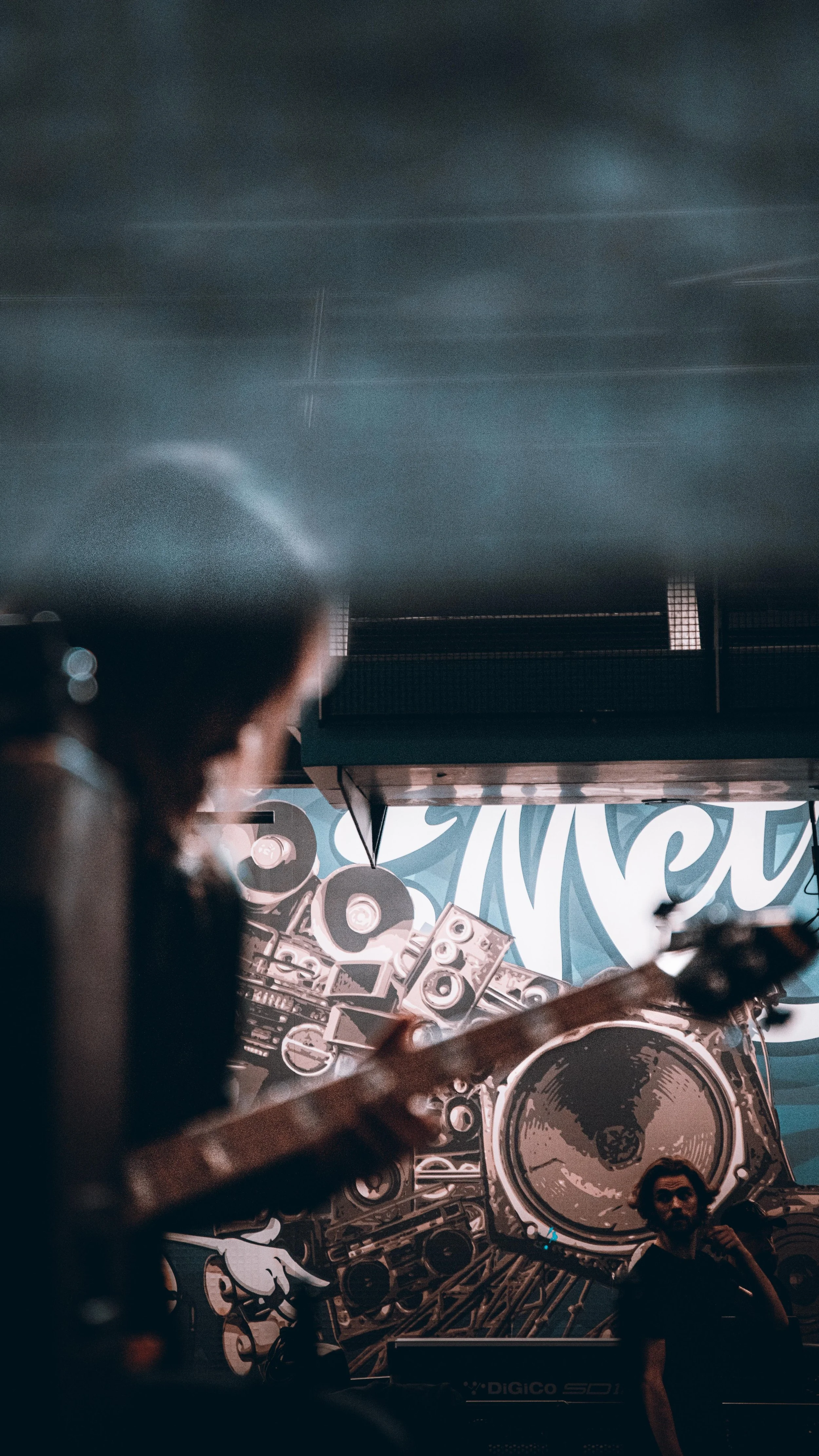 A musician playing guitar in front of a backdrop with a large speaker and musical equipment, with a person standing nearby. The scene appears to be at a live music event or concert.