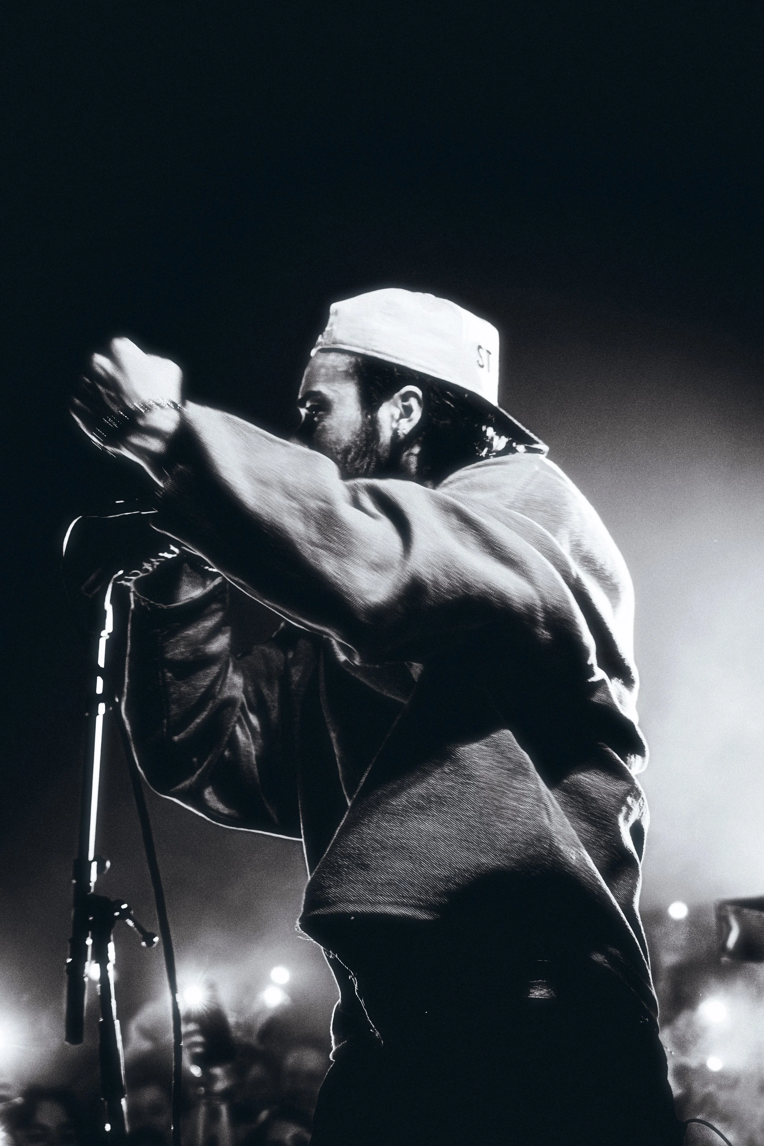 Black and white photo of a man singing or speaking into a microphone on stage, wearing a cap and a jacket.