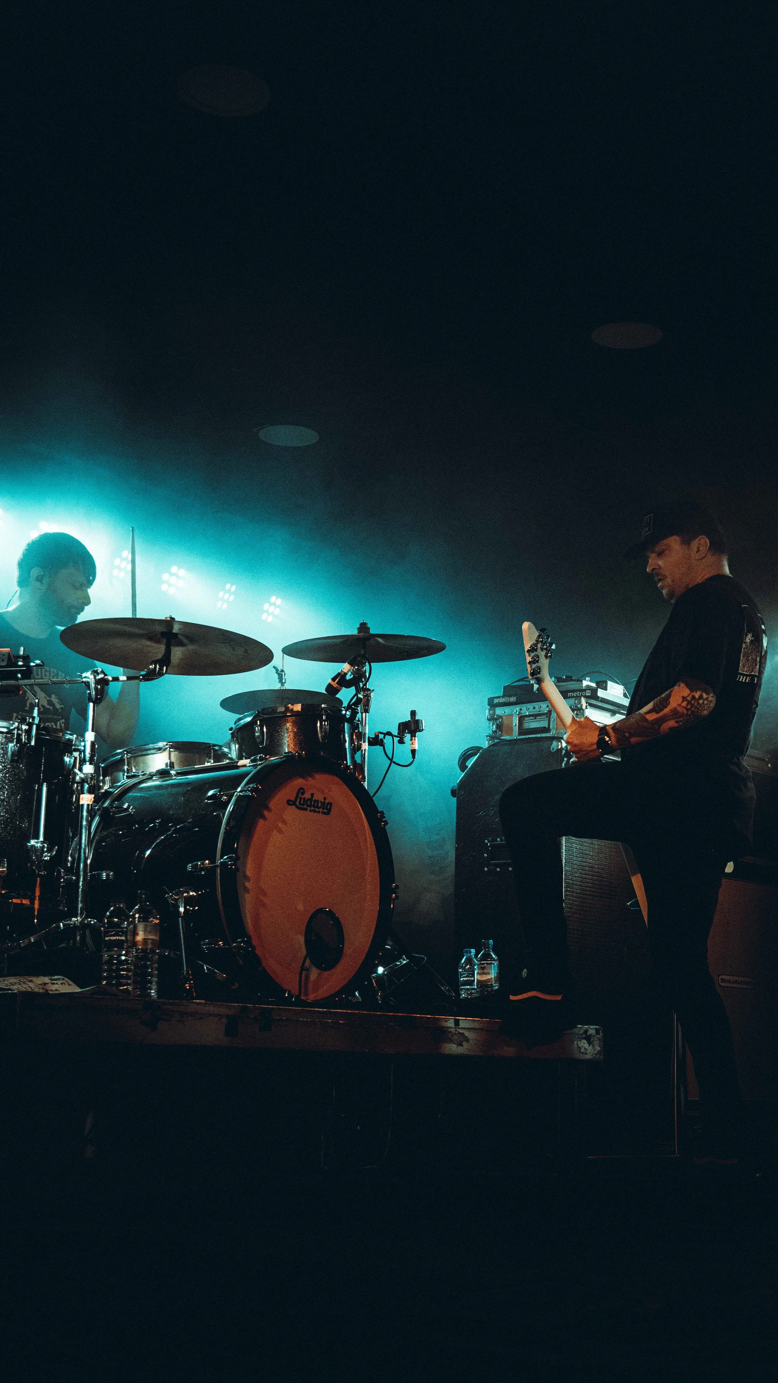 Musicians performing on a dark stage with blue lighting, one playing drums and the other playing an electric guitar, with water bottles on the floor.