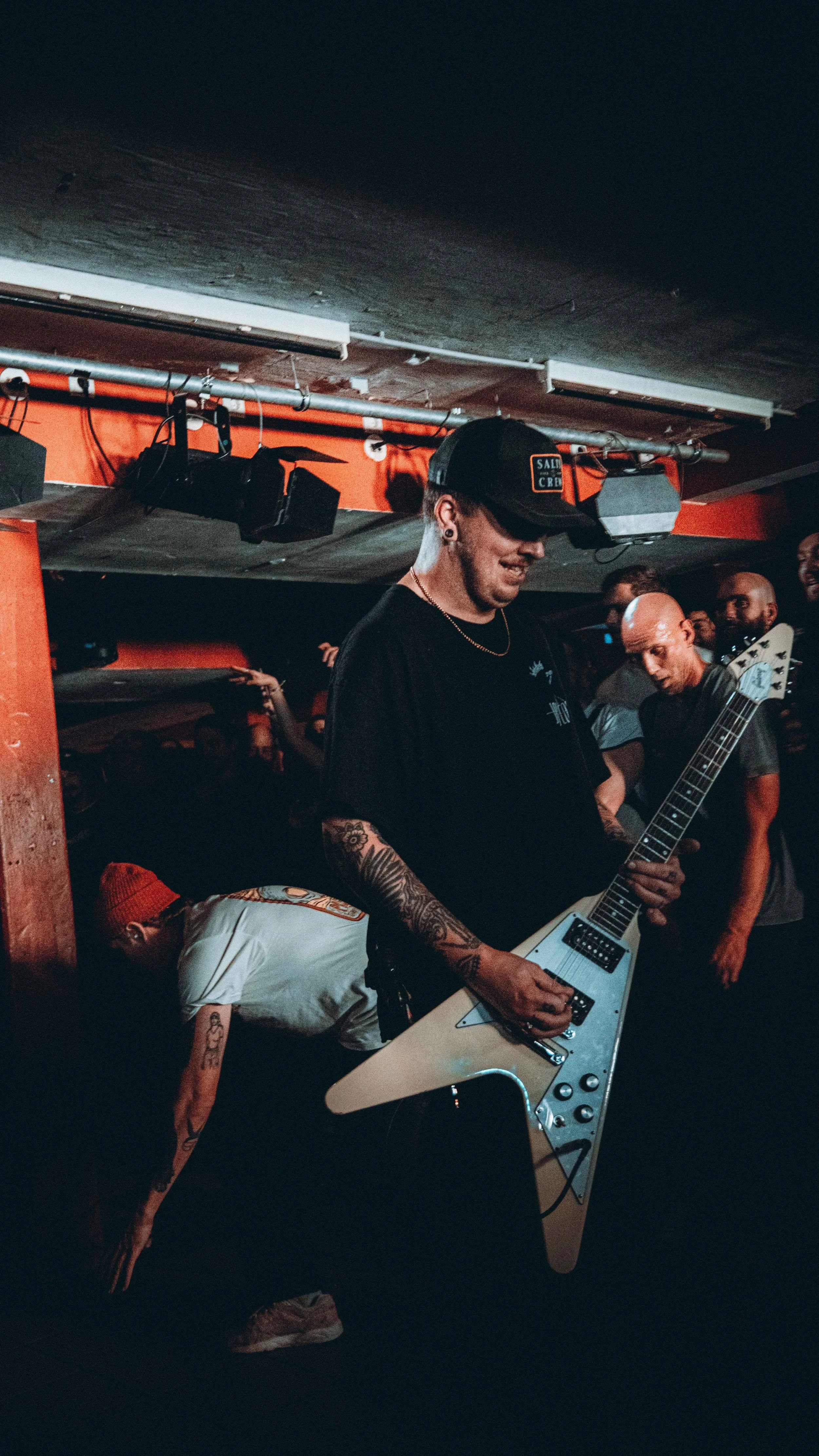 A man playing an electric guitar at a music venue with a group of people in the background.