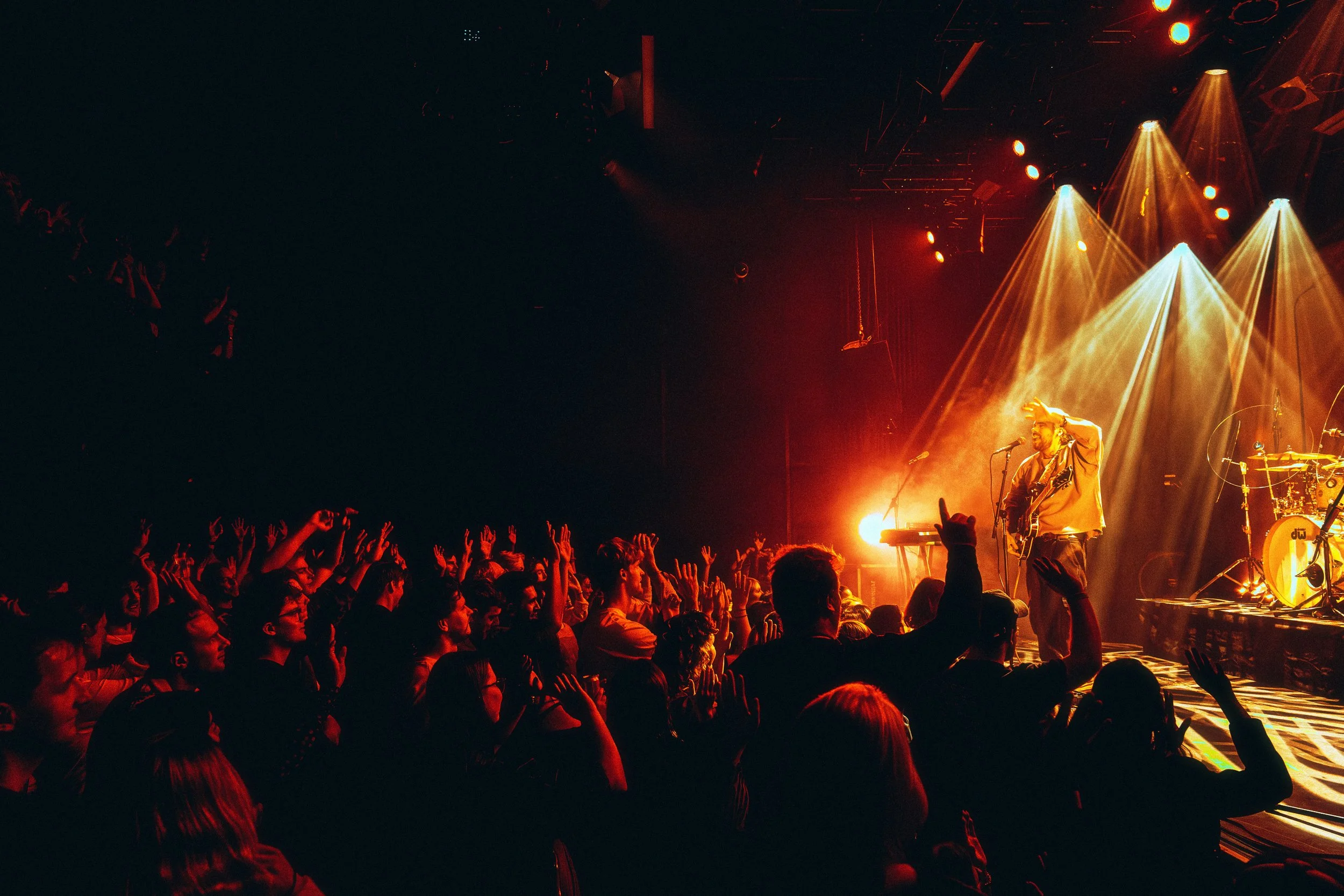 A live concert scene with a performer singing and playing guitar on stage, illuminated by warm stage lights, while the audience in the foreground reacts with raised hands and excitement.