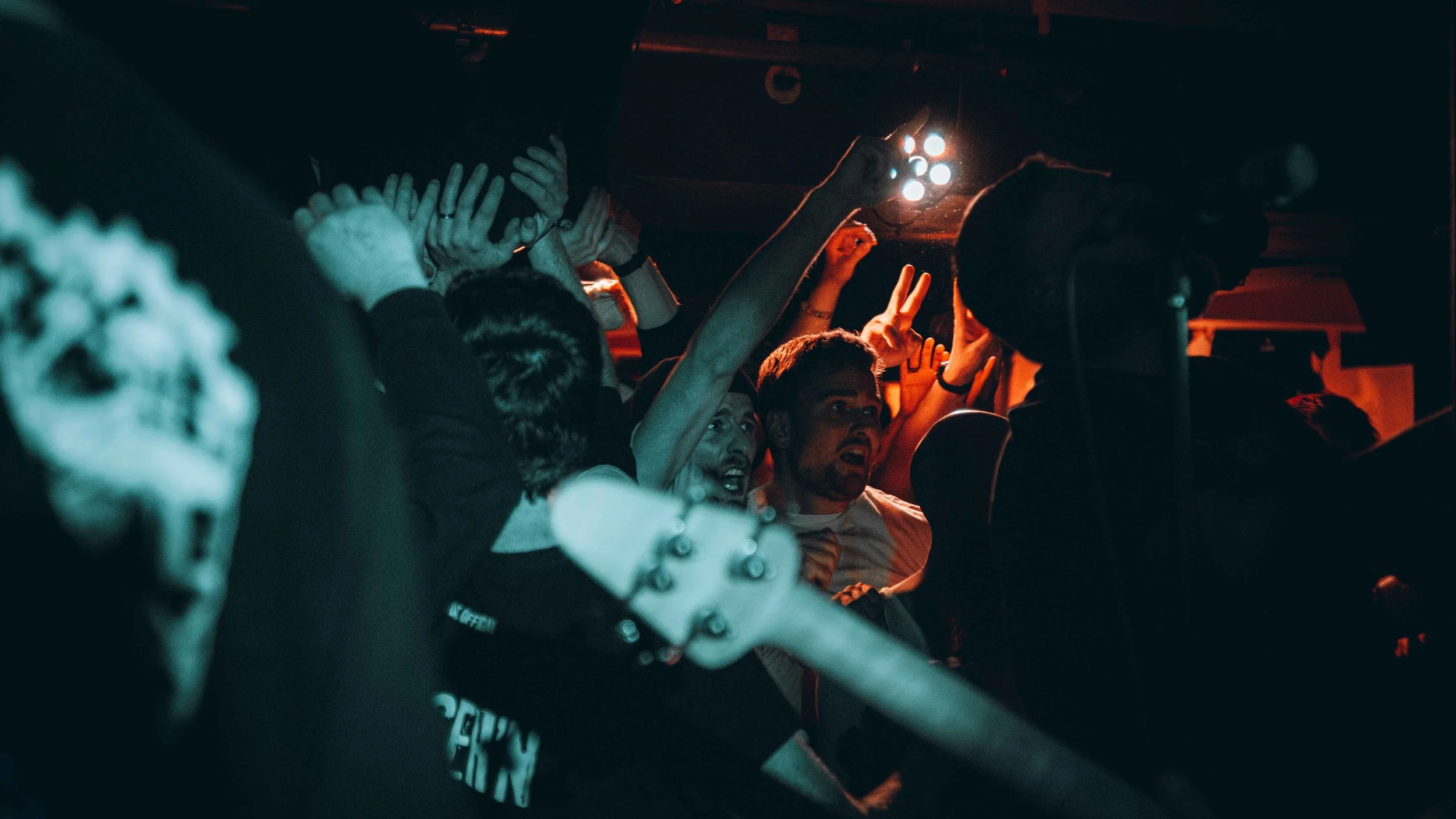 Group of people at a concert or party, some raising their hands, illuminated by stage lighting.
