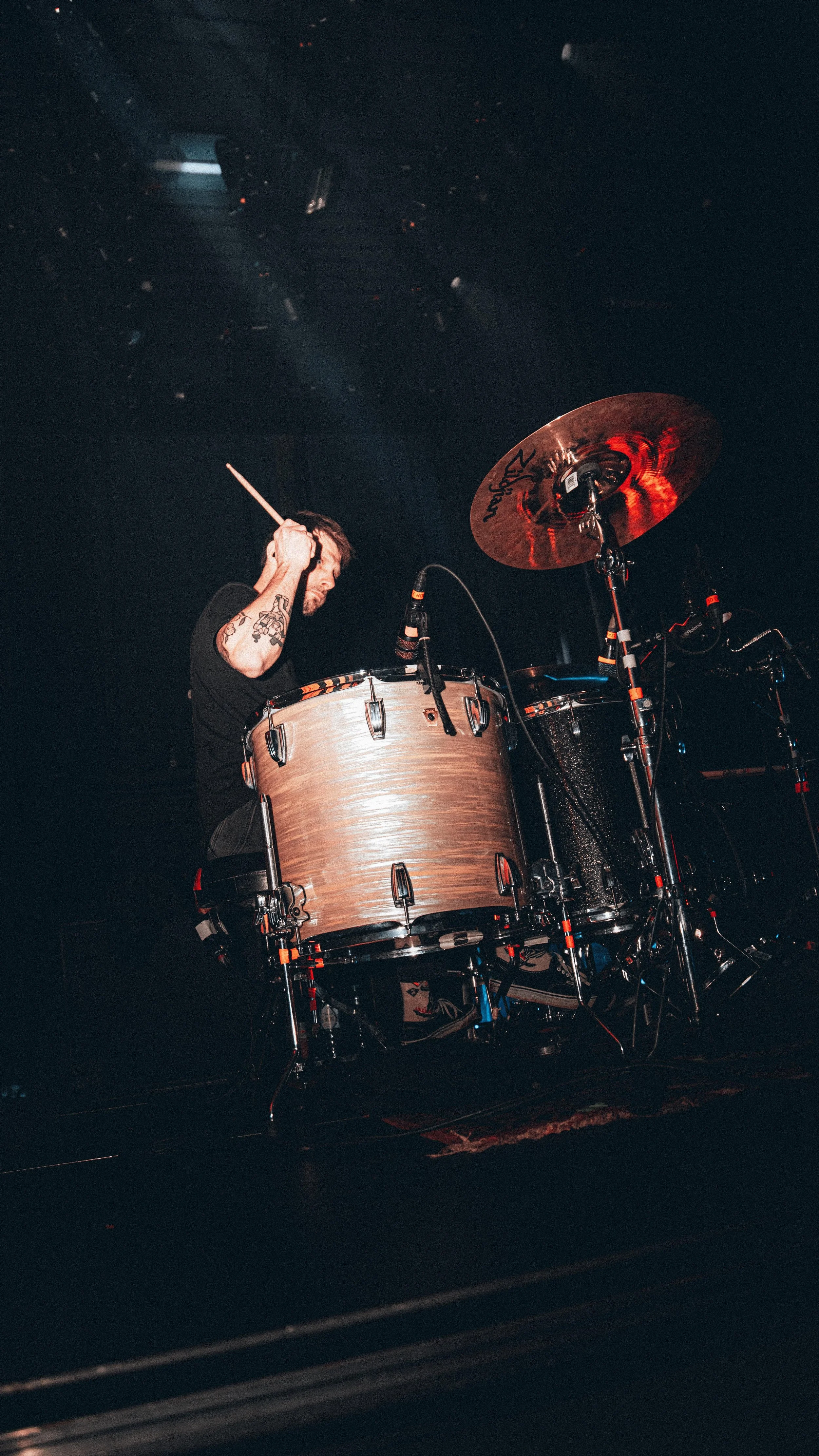A drummer with short hair and tattoos on his arm, playing a drum set on stage. The drum set includes a large bass drum and cymbals. The stage is dark with some lighting.