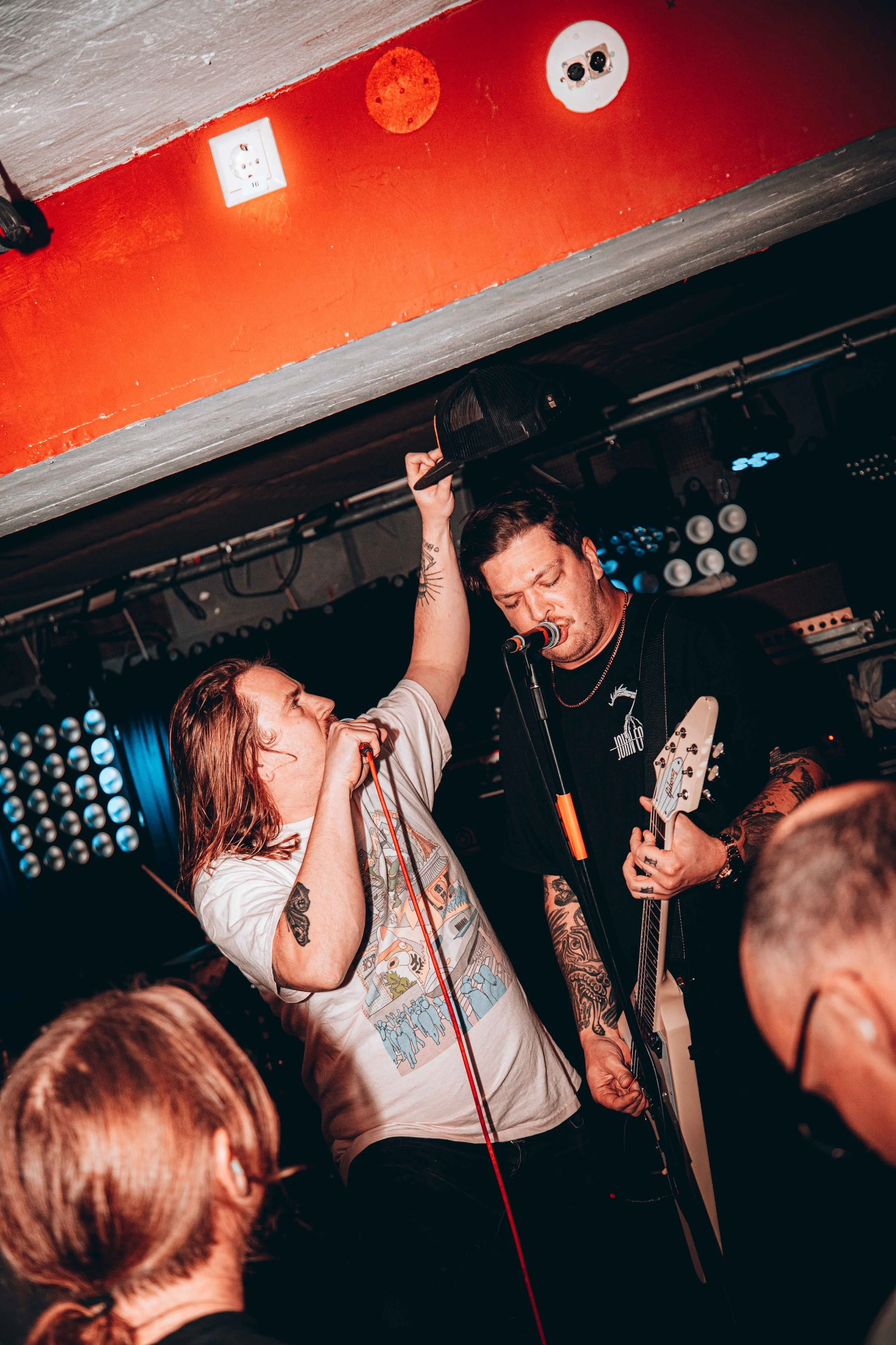 Two male musicians performing on stage with microphone and guitar, one wearing a cap, tattoos visible, audience members in foreground, stage lights and sound equipment in background.