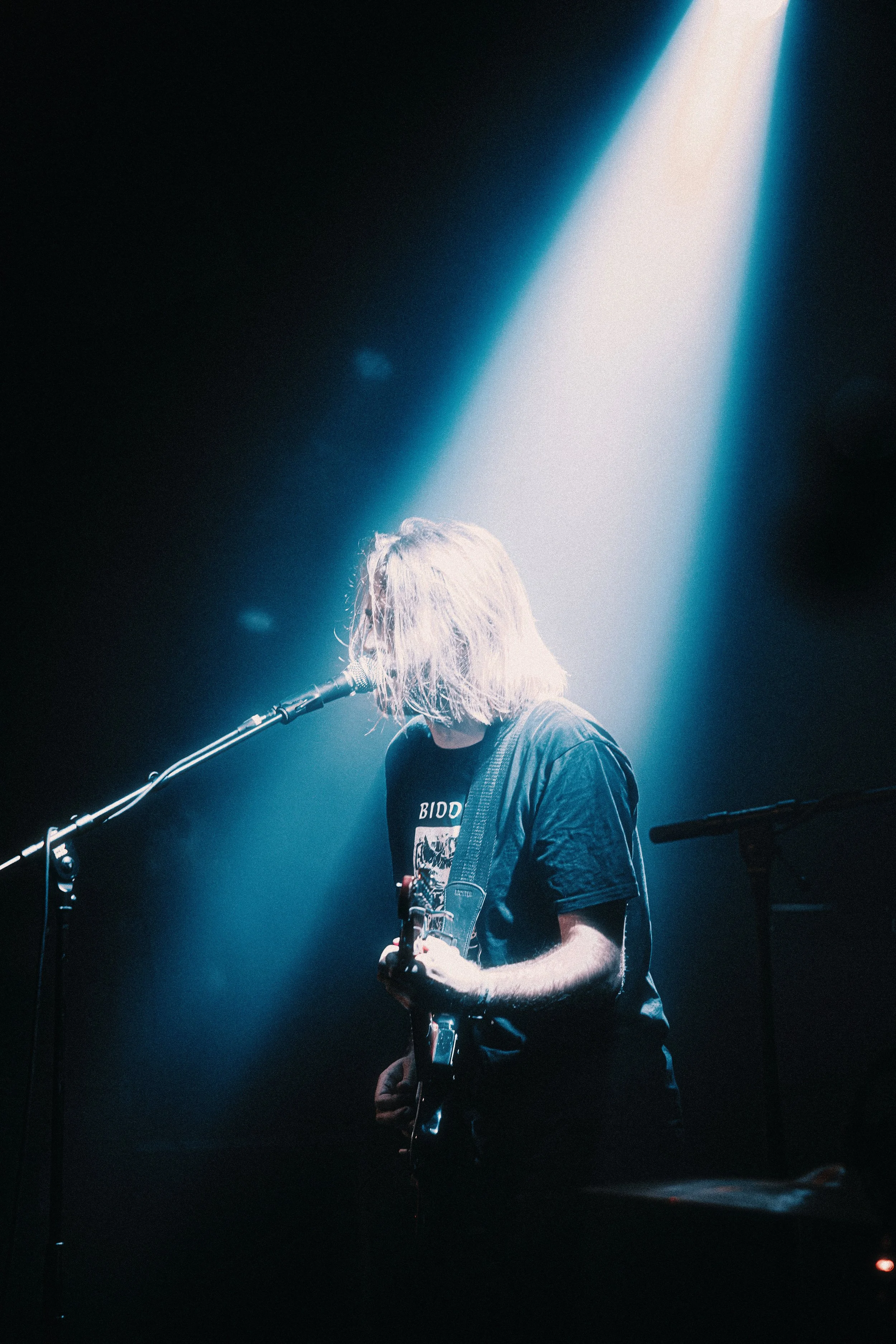 A musician with long hair performing on stage, playing an electric guitar under a bright spotlight in a dark setting.