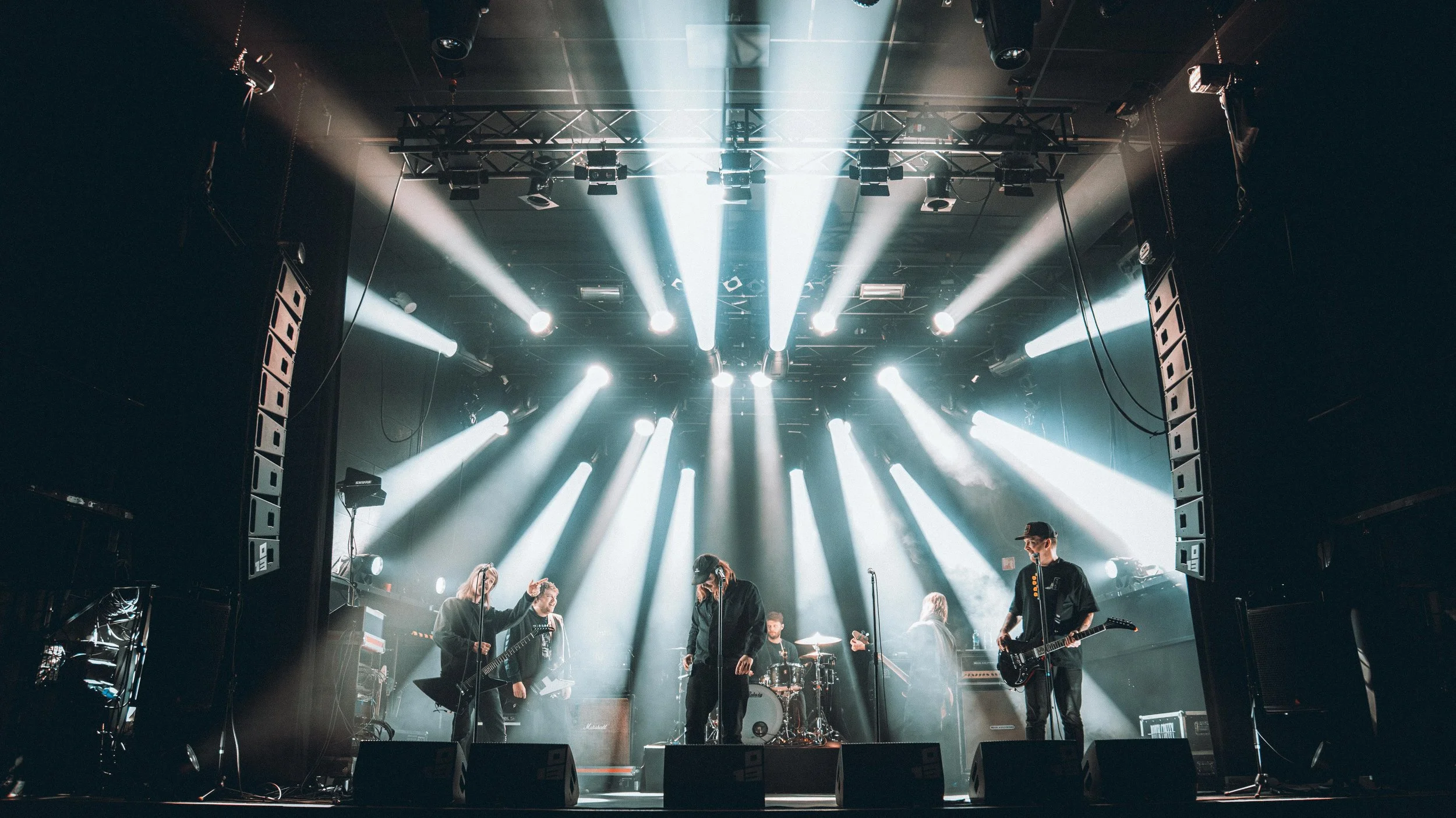 A band performing on stage with bright stage lights shining down, featuring guitarists, a drummer, and singers.