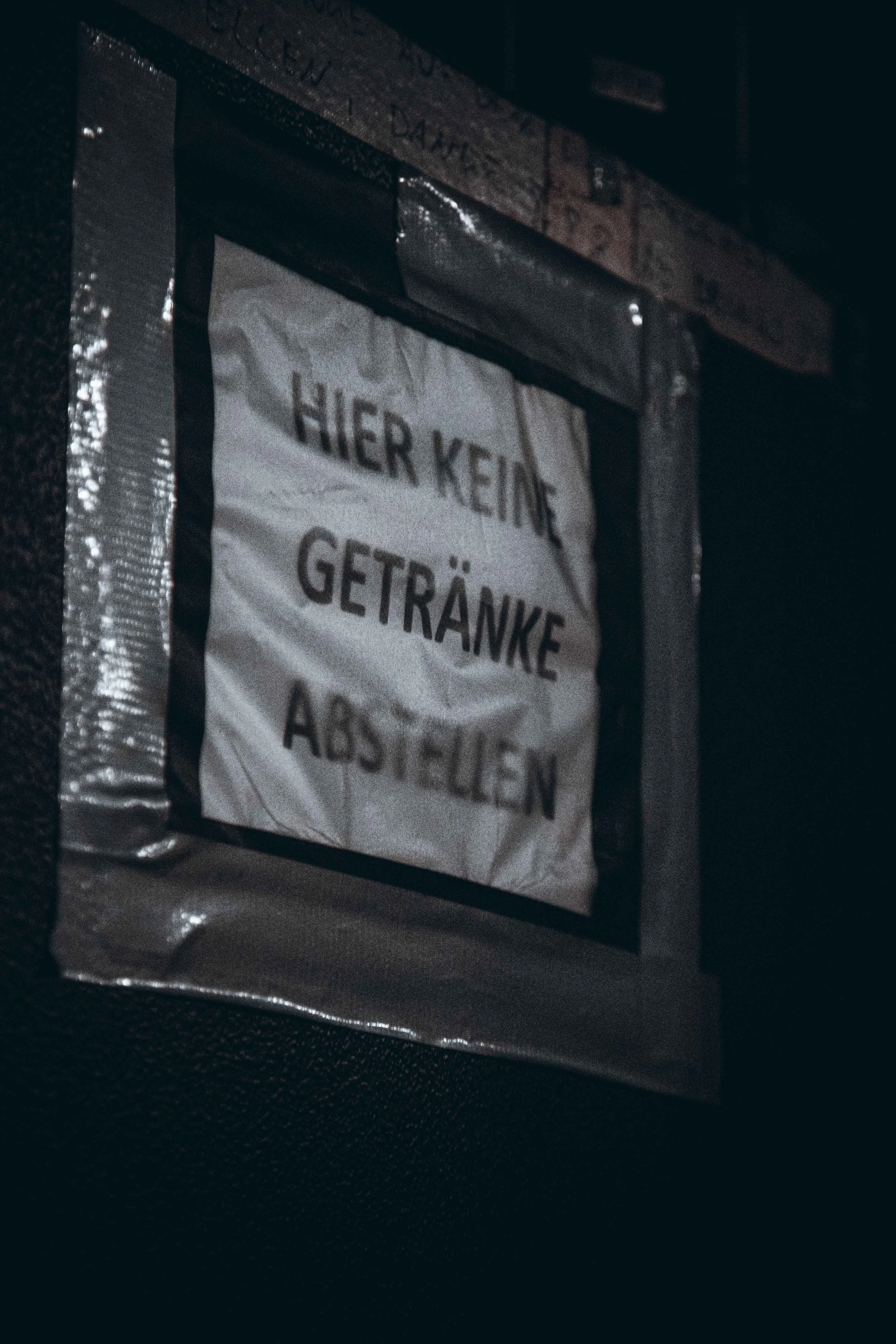 A sign with German text that reads 'HIER KEINE GETRÄNKE ABSTELLEN', taped to a dark wall.
