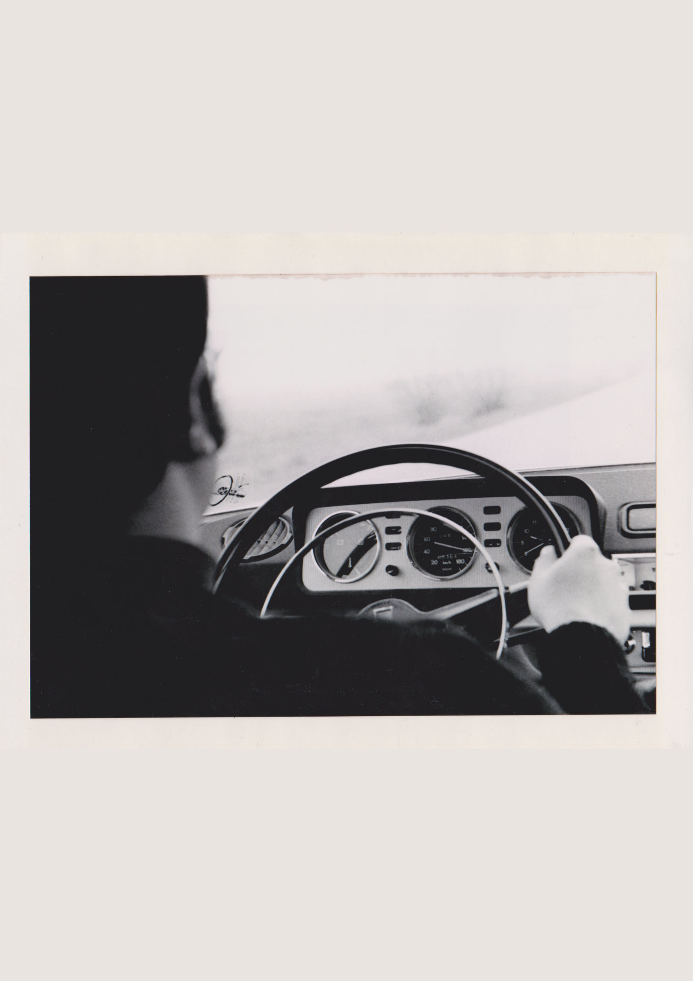 Black and white photo of a person driving a car, viewing the dashboard and steering wheel, with a blurry landscape visible through the windshield.