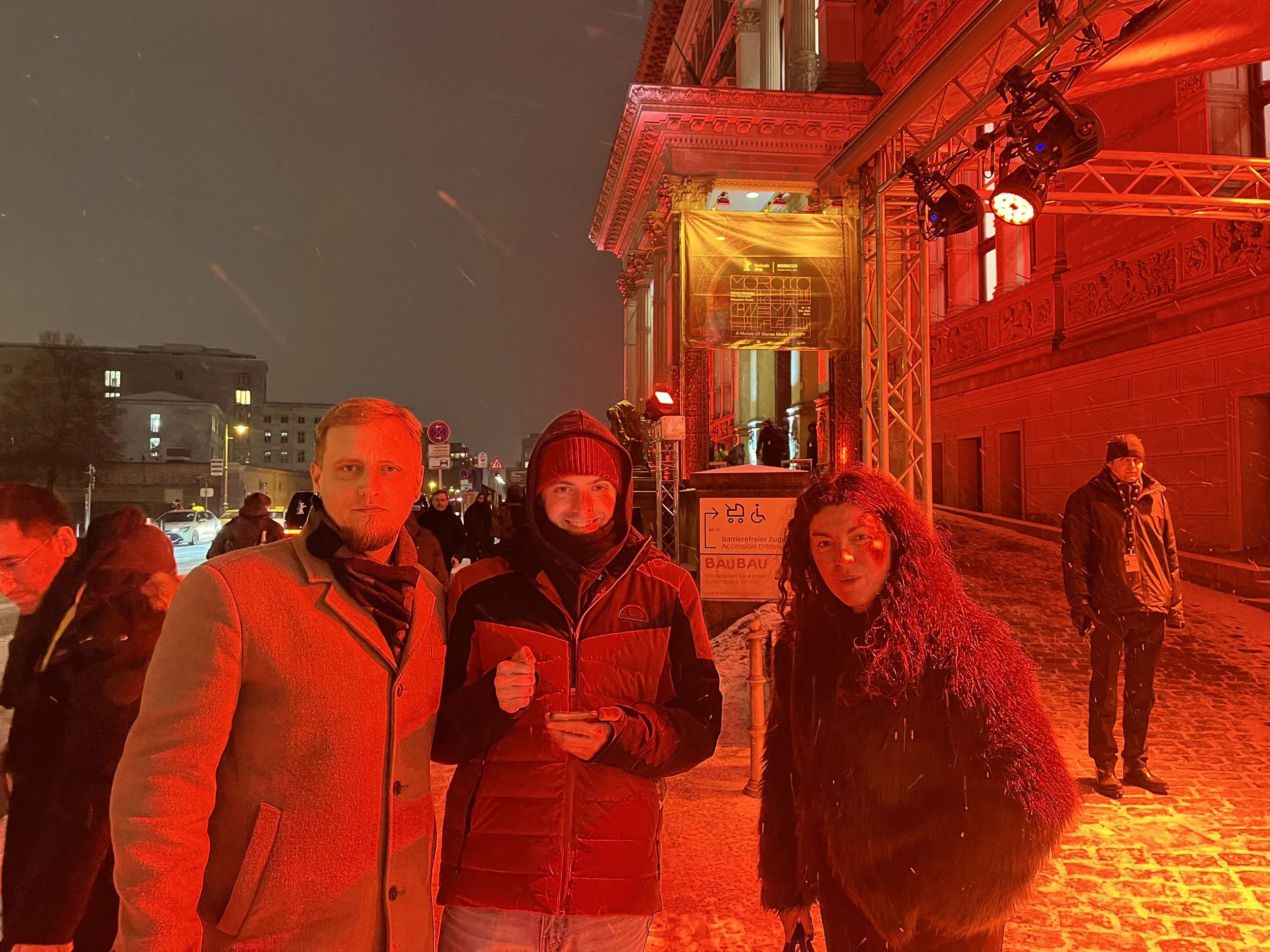Nighttime scene in front of an ornate building illuminated in red light, with a group of people posing for a photo, including a man with a beard, a man in a beanie, and a woman with curly hair.