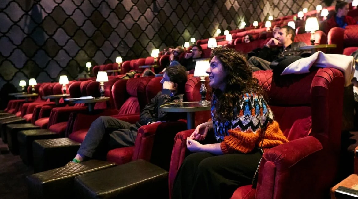 People watching a movie in a theater with red velvet seats and small lamps on each table.
