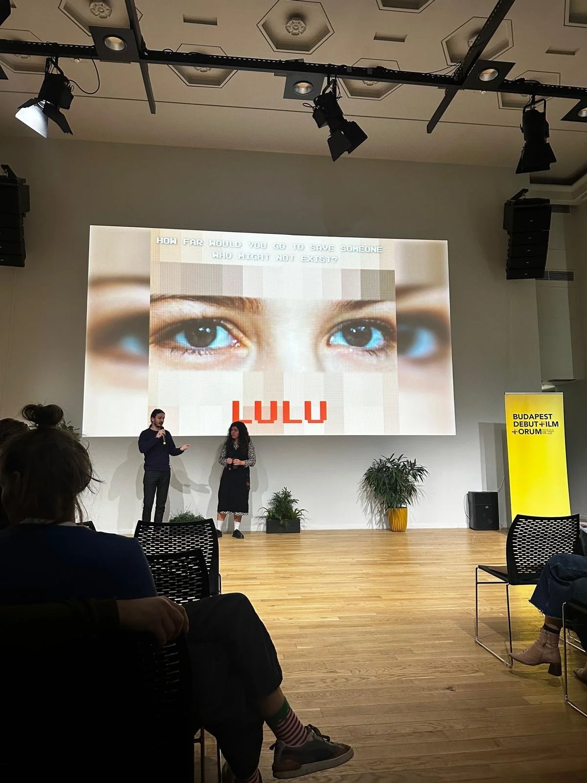 A presentation in a conference room with two speakers on stage, a large screen displaying a close-up of a child's eyes and the text 'HOW FAR WOULD YOU GO TO SAVE SOMEONE WHO MIGHT NOT EXIST?' and 'LULU'. There are potted plants on stage, audience mem