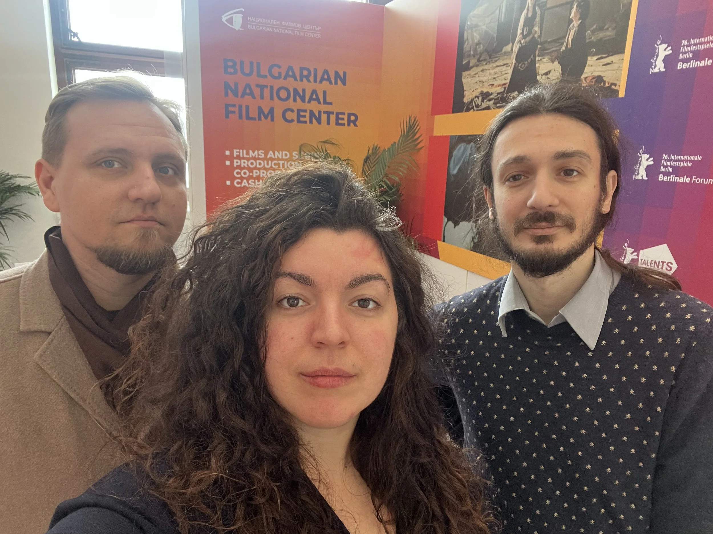 Three people taking a selfie in front of a colorful poster promoting the Bulgarian National Film Center and the Berlinale film festival, with a woman in the center and two men on either side.