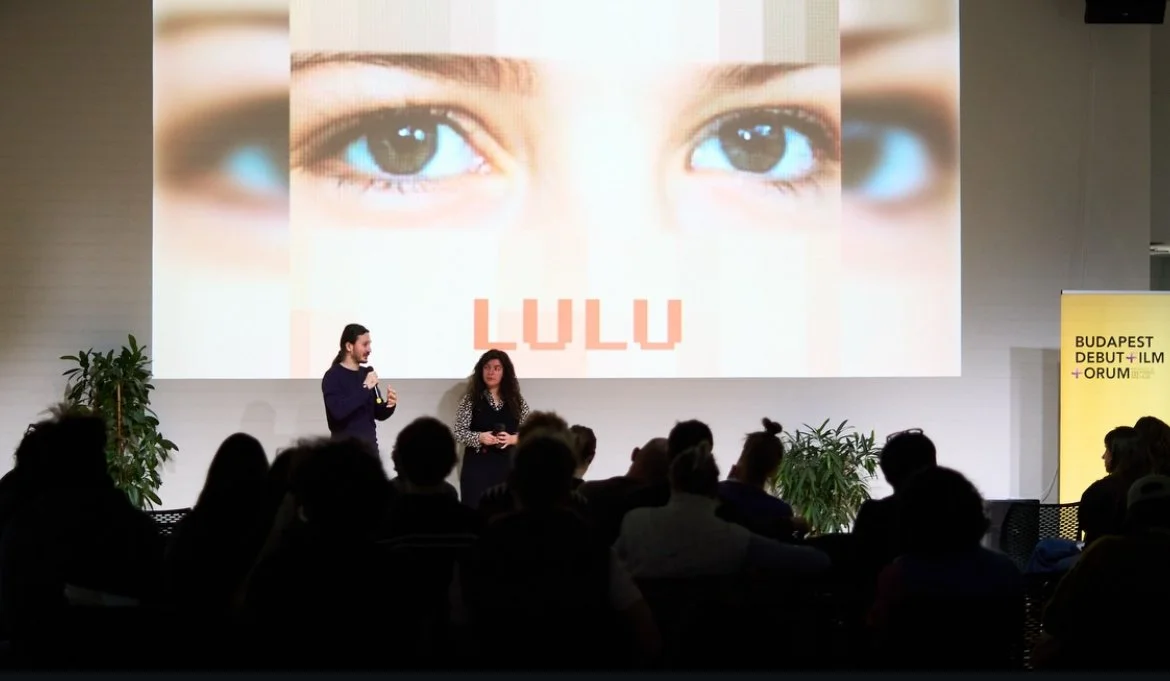 A presentation at an event with a large screen displaying a close-up of a child's eyes and the text 'LULU'. Two presenters stand in front of an audience, with potted plants nearby and a yellow banner on the side that reads 'BUDAPEST DEBUT + ILM + ORU