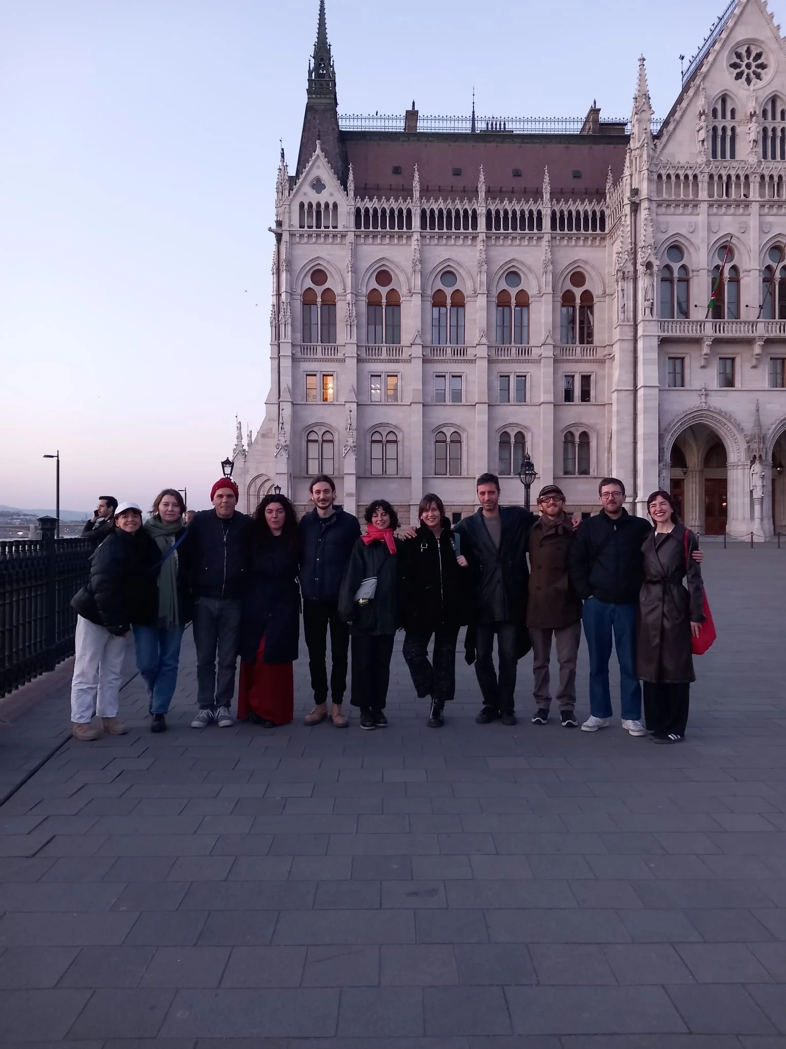 A group of people standing outdoors in front of a historic, gothic-style building with spires and ornate windows, during twilight or early evening.