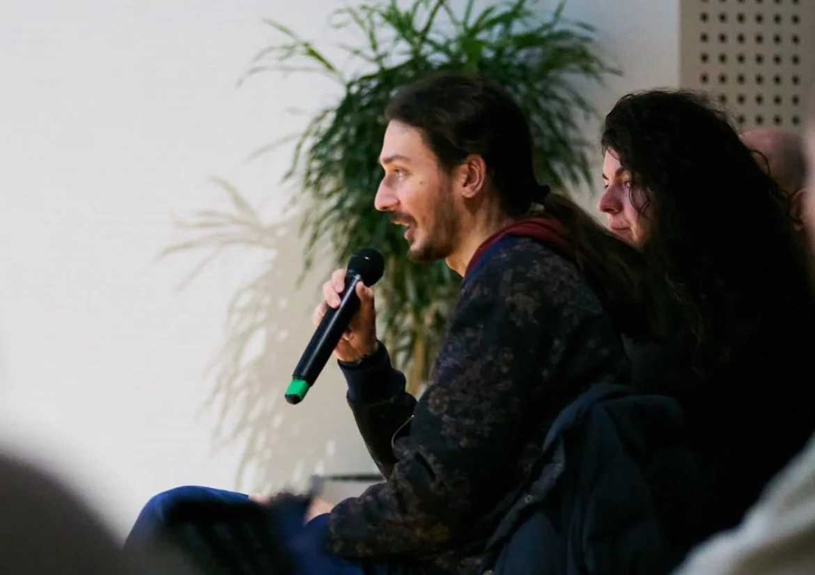 A man with long dark hair and a beard speaking into a microphone during a panel discussion or interview at an indoor event, with a woman sitting beside him and a potted plant in the background.