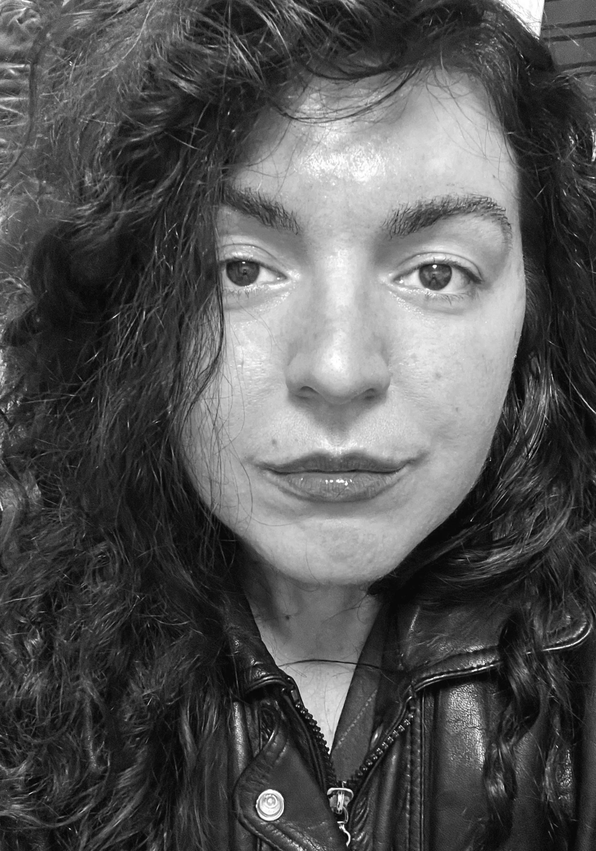 Close-up of a woman with curly hair, wearing a leather jacket, looking at the camera with a neutral expression.