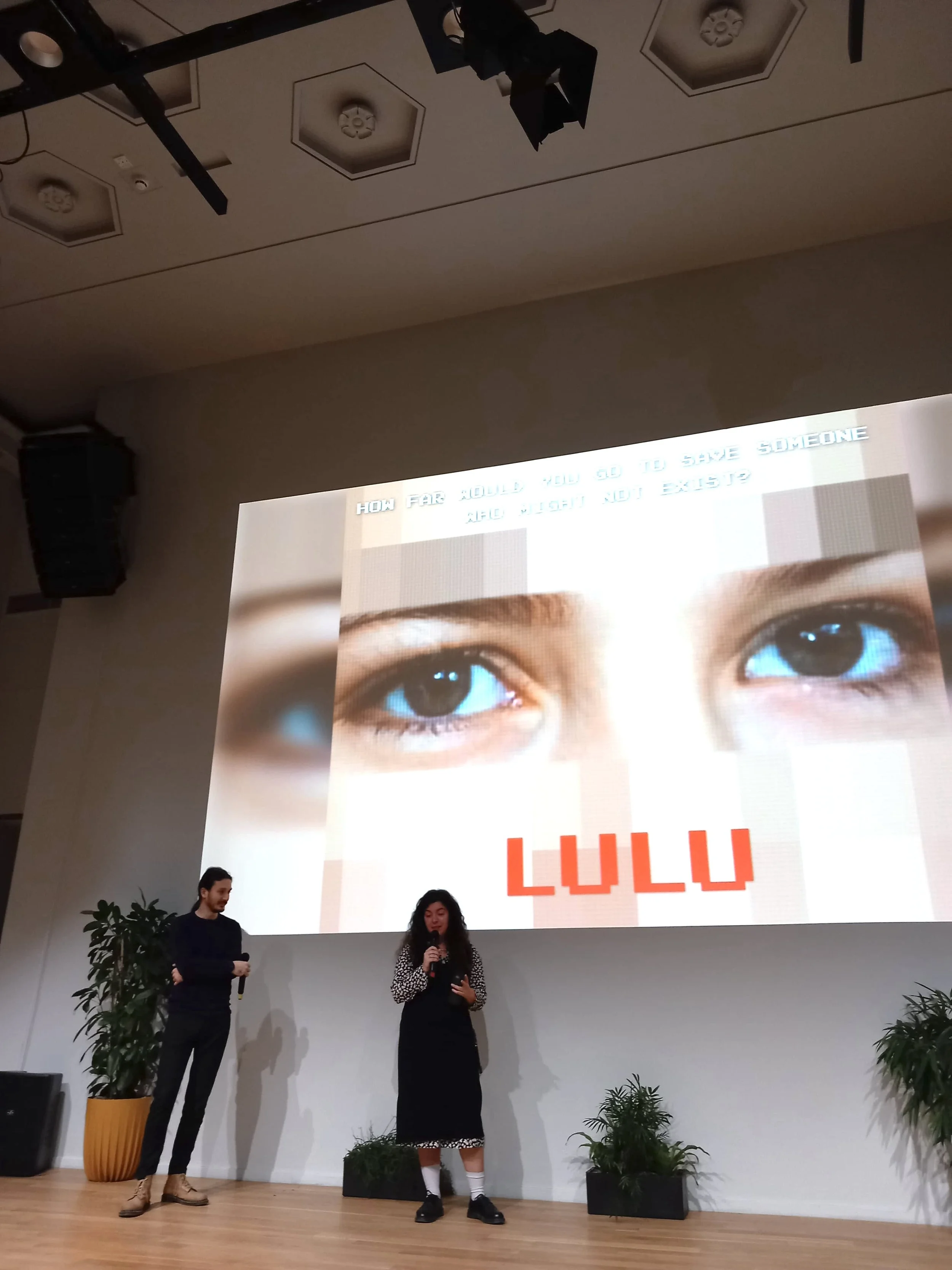 A woman and a man standing on a stage in front of a large screen displaying an image of eyes and the word 'LULU'. The woman is speaking into a microphone. There are plants on either side of the stage, and the audience is not visible.