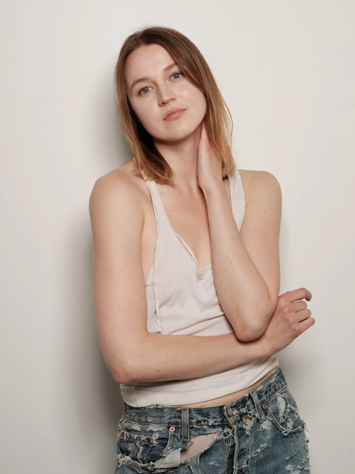 Jeune femme aux cheveux roux, portant un t-shirt blanc déchiré et un short en jean troué, posant contre un mur blanc.