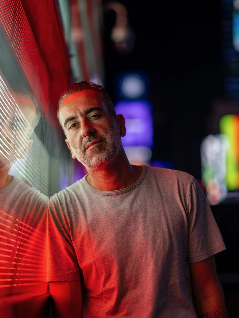 Un homme avec une barbe, portant un t-shirt gris clair, avec un fond de lumières de néon colorées, dans un environnement nocturne urbain.