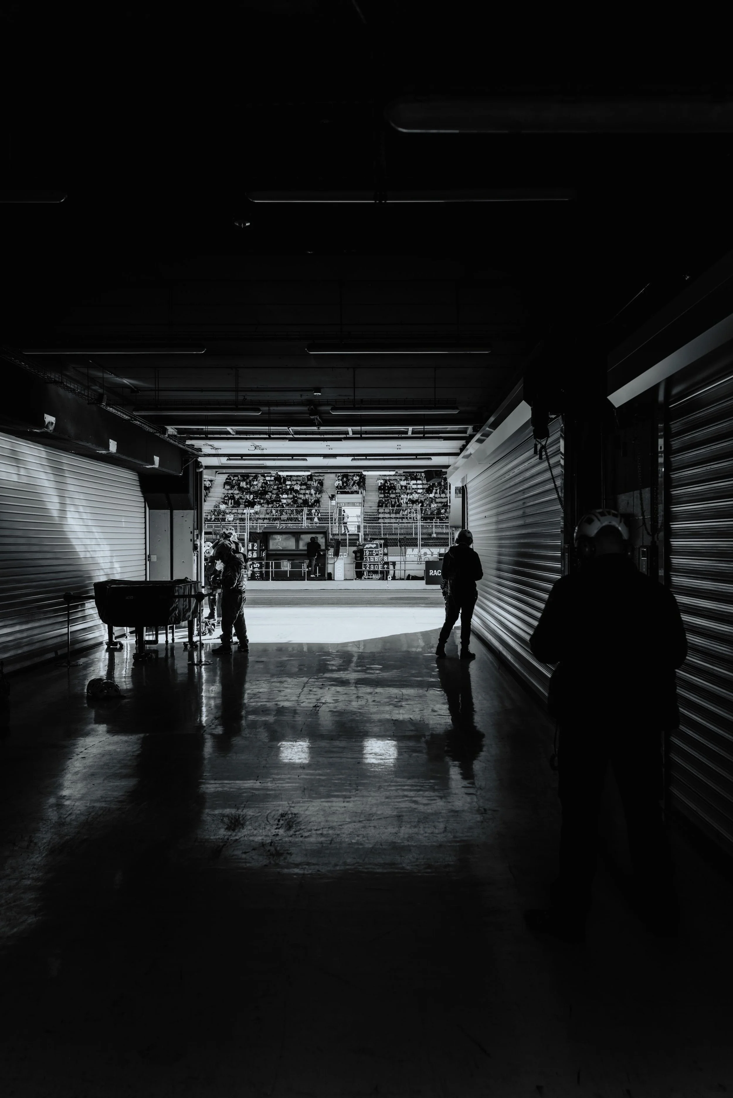 Une scène d'intérieur en noir et blanc d'une zone du circuit de course, avec des personnes debout près des roulottes fermées et une vue de la piste de course en arrière-plan, où se trouvent des spectateurs.