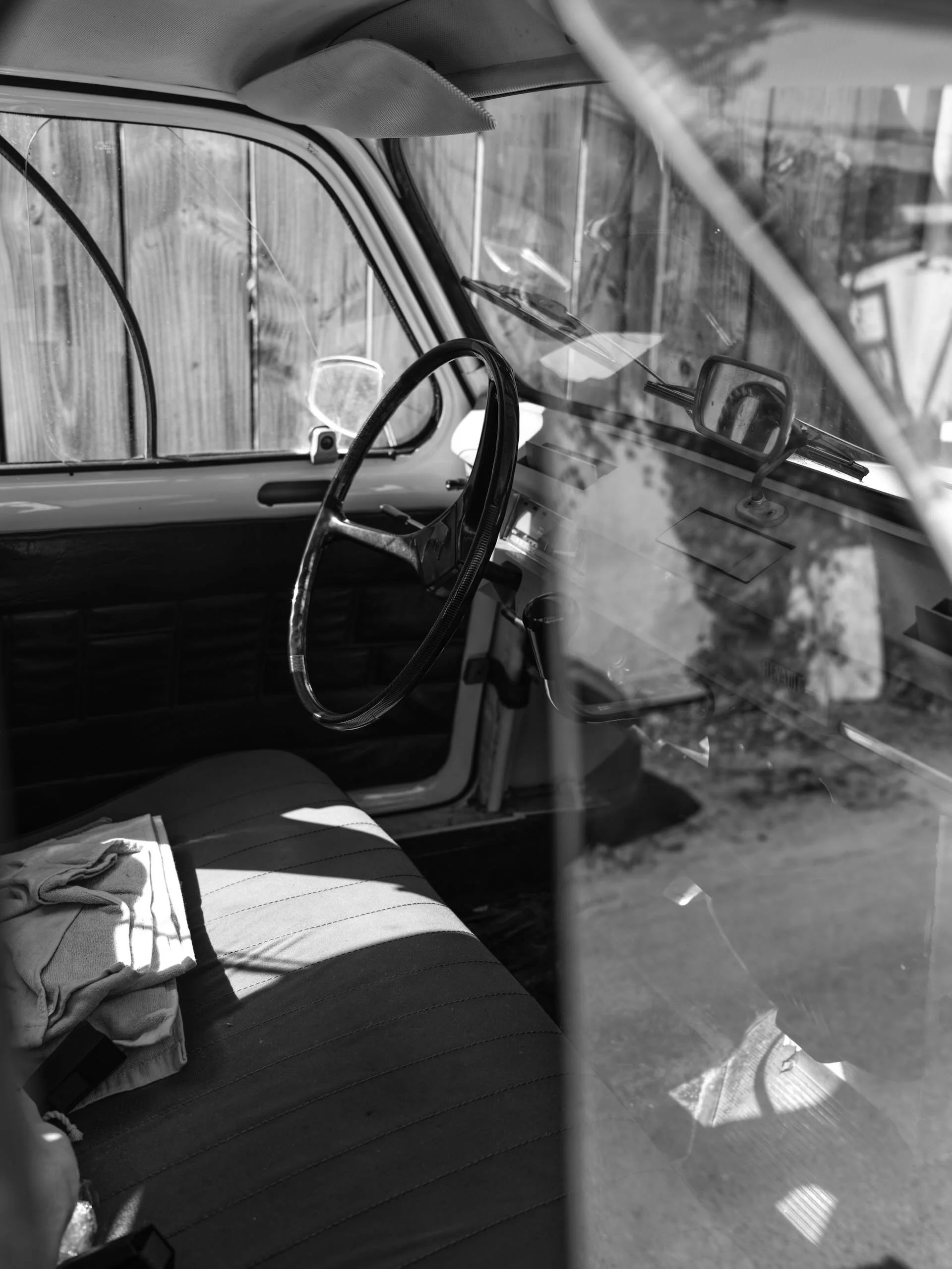 Intérieur d'une vieille voiture en noir et blanc, vue du conducteur, avec un volant, un tableau de bord, un rétroviseur, et un siège avec des vêtements. La porte est ouverte et une clôture en bois est visible à l'extérieur.