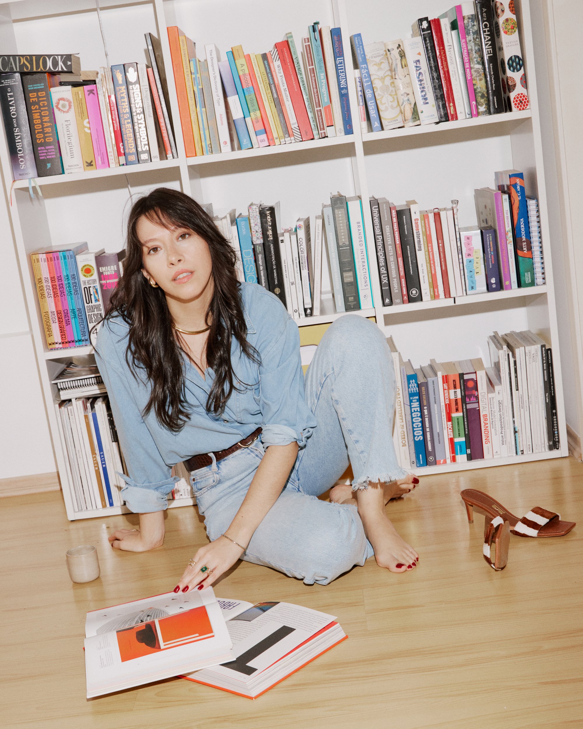 Mulher de cabelos longos e escuros sentada no chão de frente para a câmera, com uma livros abertos na frente, ao lado de uma vela, usando jeans e uma camisa azul, ao lado de prateleiras cheias de livros.