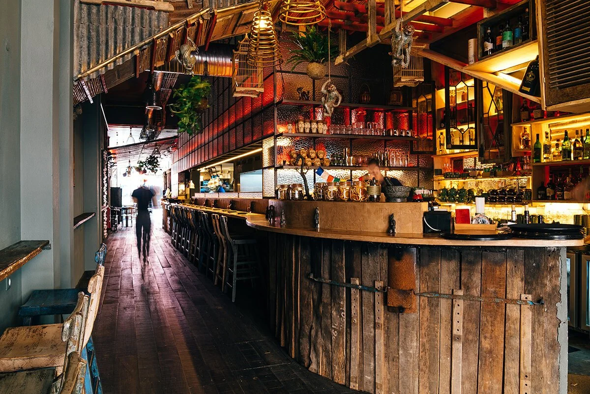 Interior of a bar with rustic wooden decor, barstools lined along the wooden counter, shelves stocked with bottles, and dim ambient lighting. A person is walking away towards the back of the establishment.