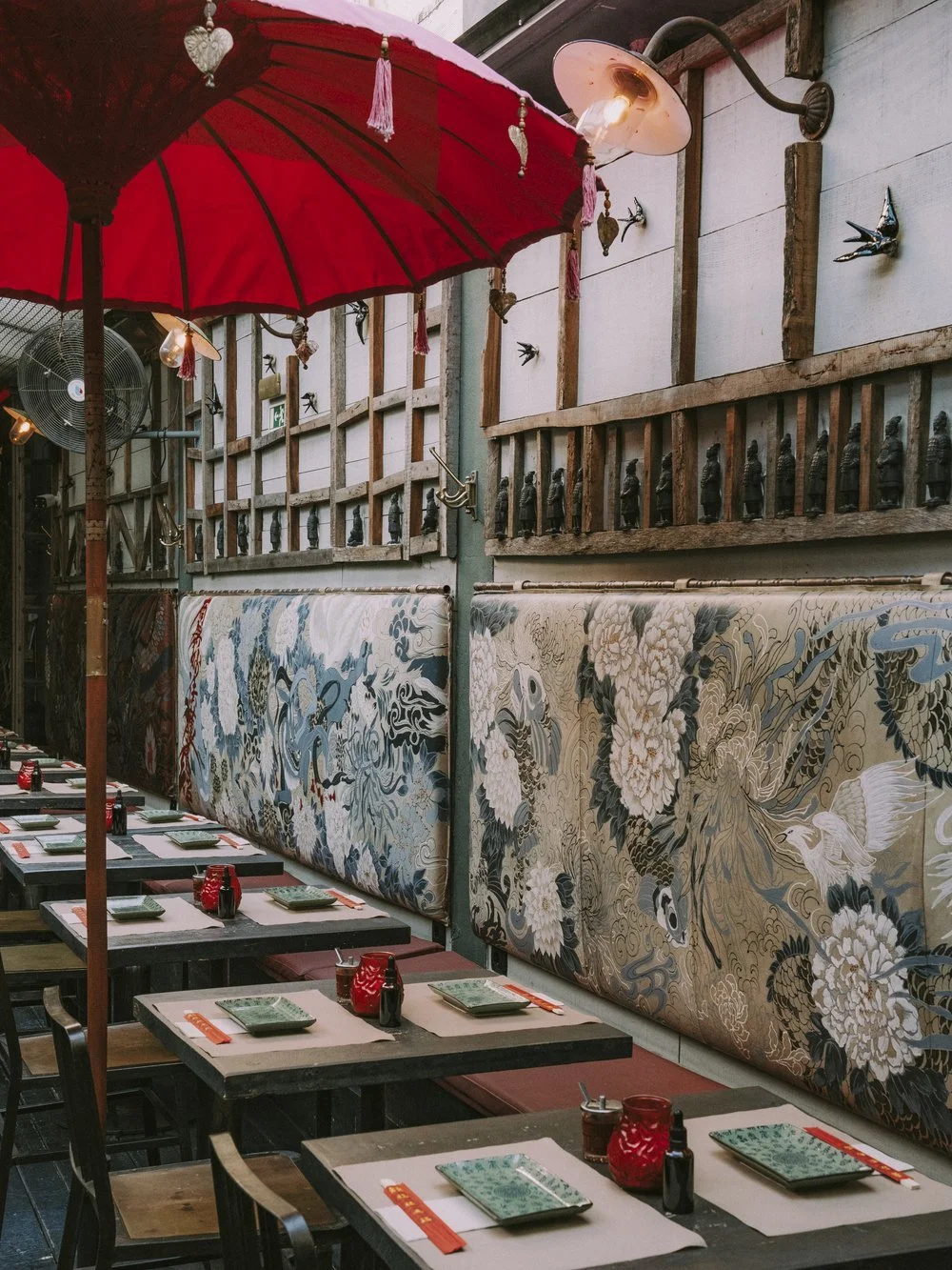 Outdoor restaurant seating area with tables set with plates, chopsticks, and condiments, shaded by a large red umbrella, and decorated with Asian-inspired wall art and hanging ornaments.
