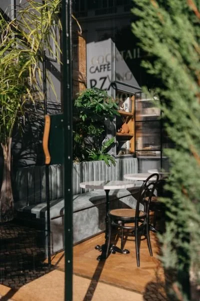 Outdoor patio area with a small round table and two black chairs, lush greenery, and a cafe sign in the background.
