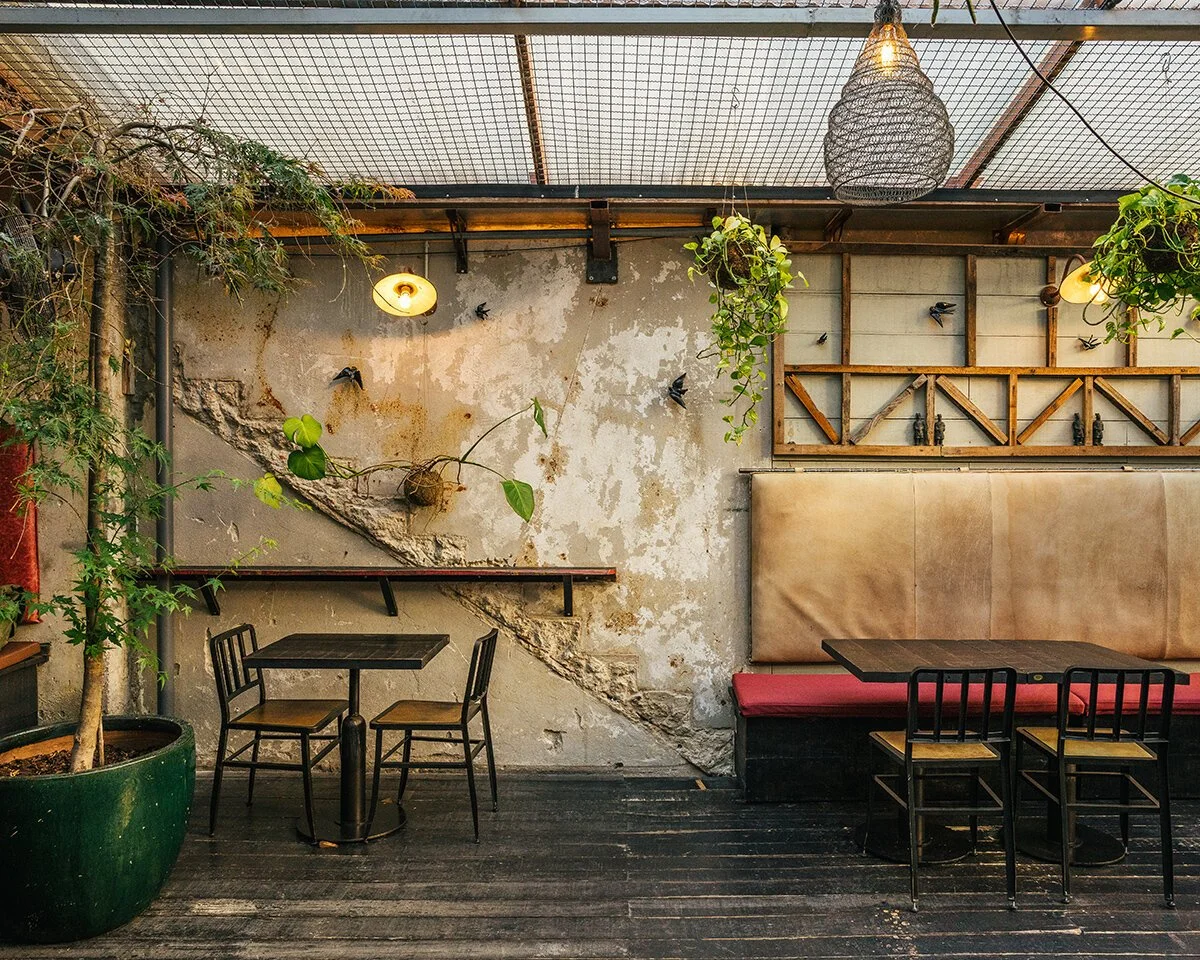 Interior of a cafe or restaurant with rustic and industrial decor, including a potted tree, hanging plants, a textured wall with peeling paint, black chairs, and tables, pendant lighting, and a cushioned booth.