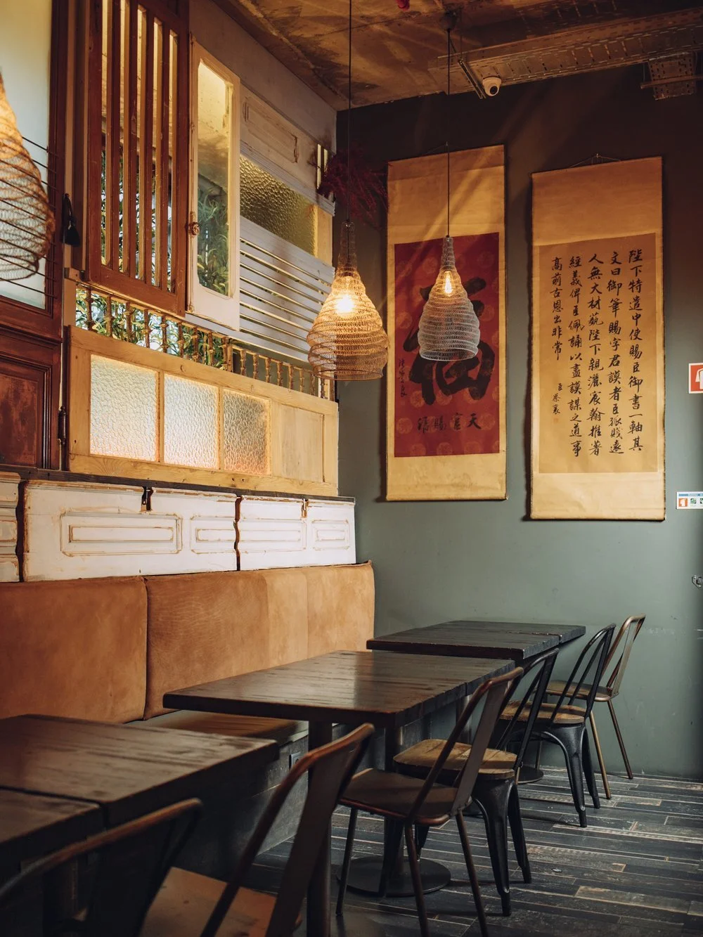 Interior of a cozy cafe with wooden tables, black and metal chairs, a cushioned bench, hanging woven pendant lights, and traditional Chinese wall scrolls.