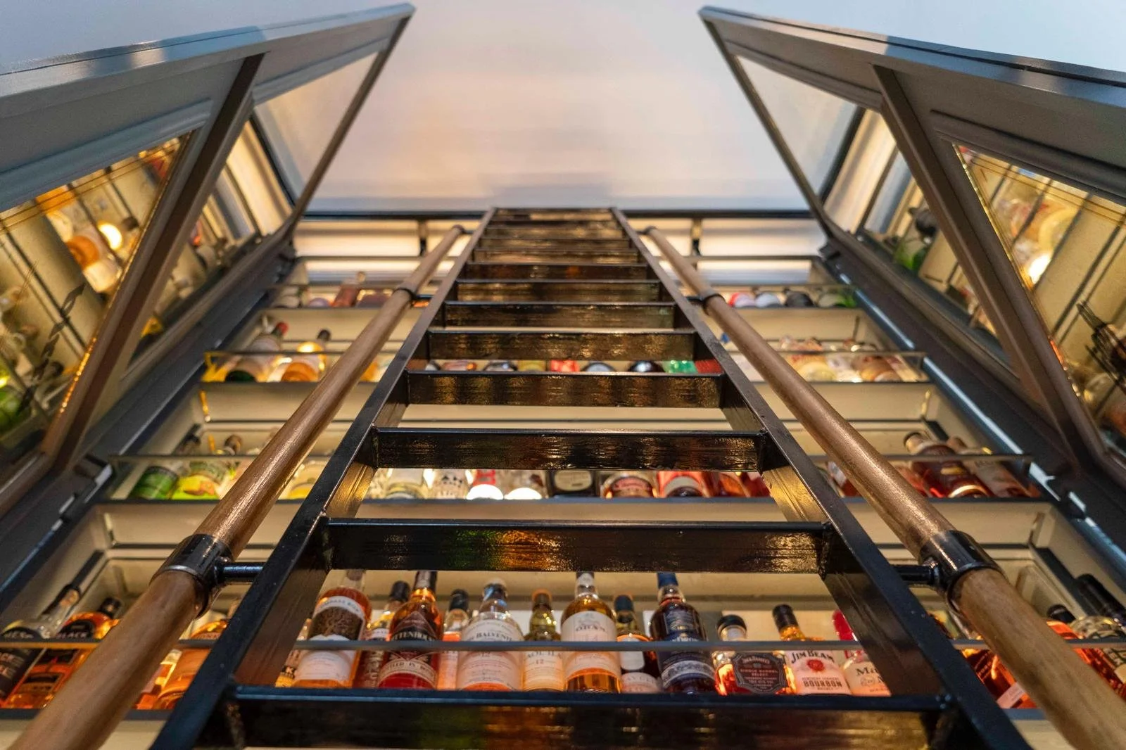 Inside view of a liquor store with a tall shelving unit filled with bottles, viewed from the bottom looking up, with a ladder in the center.