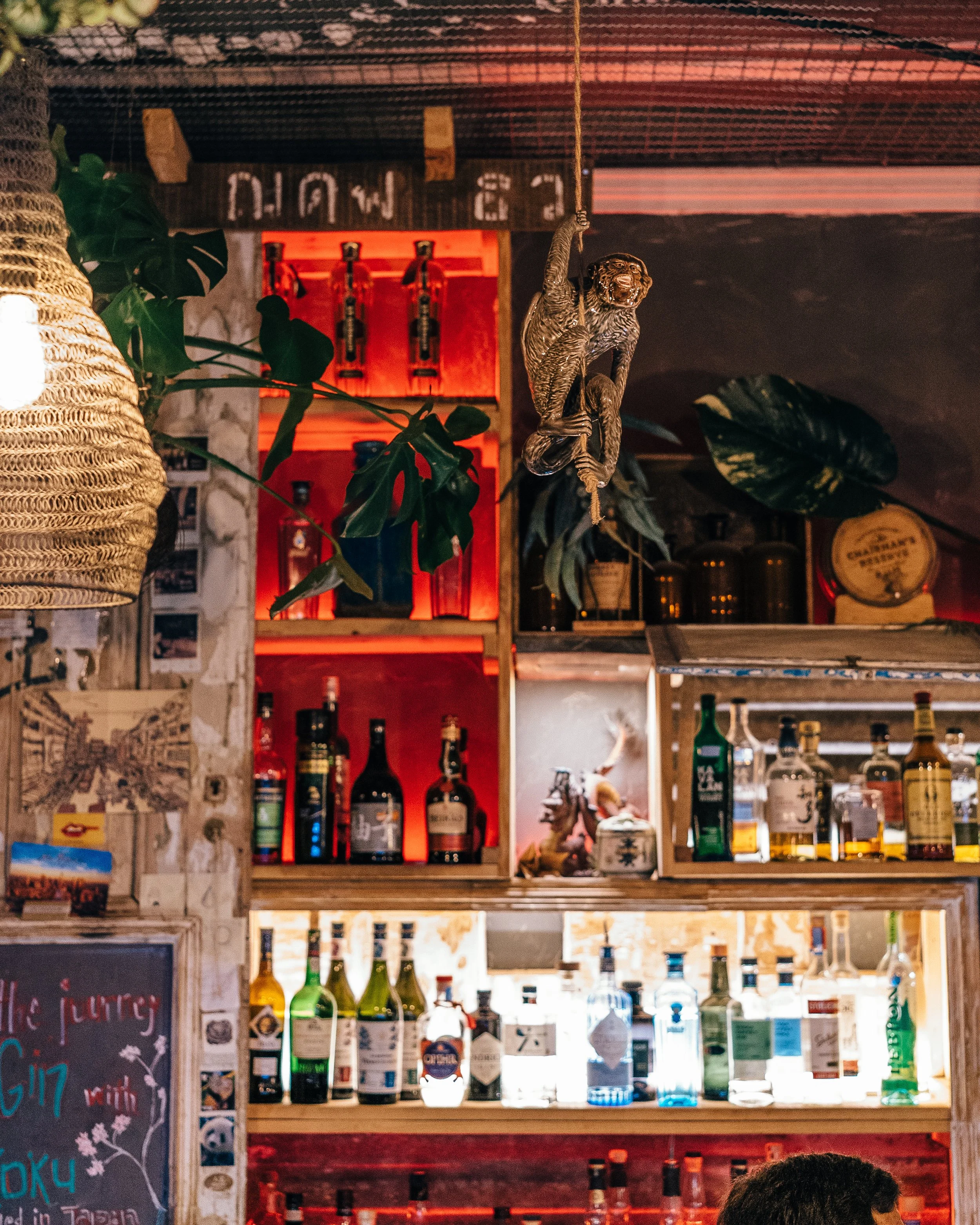 Interior of a bar with shelves of liquor bottles, a monkey figurine hanging from the ceiling, and decorative plants.