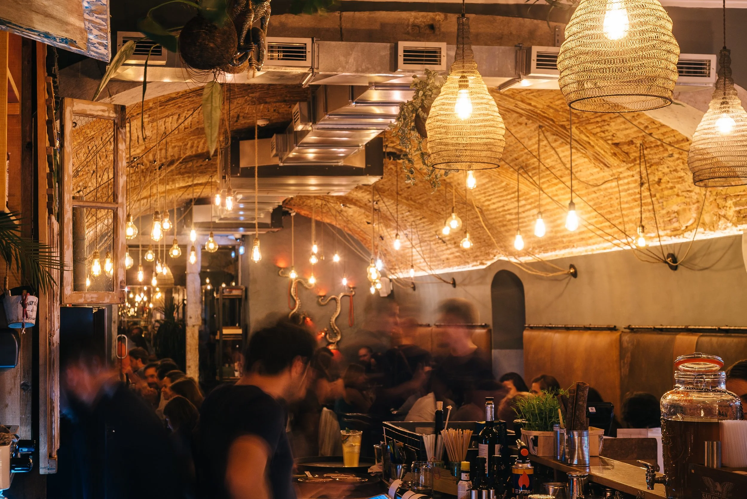 Interior of a busy restaurant or bar with warm lighting, hanging pendant lights, exposed brick ceiling, and several patrons seated at tables and the bar. Decor includes plants and wall art.