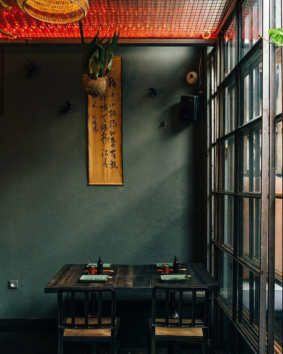 A small dining table set for two in a cozy restaurant with dark walls, a hanging plant, and a Japanese calligraphy scroll. Glass windows on the right let in natural light.