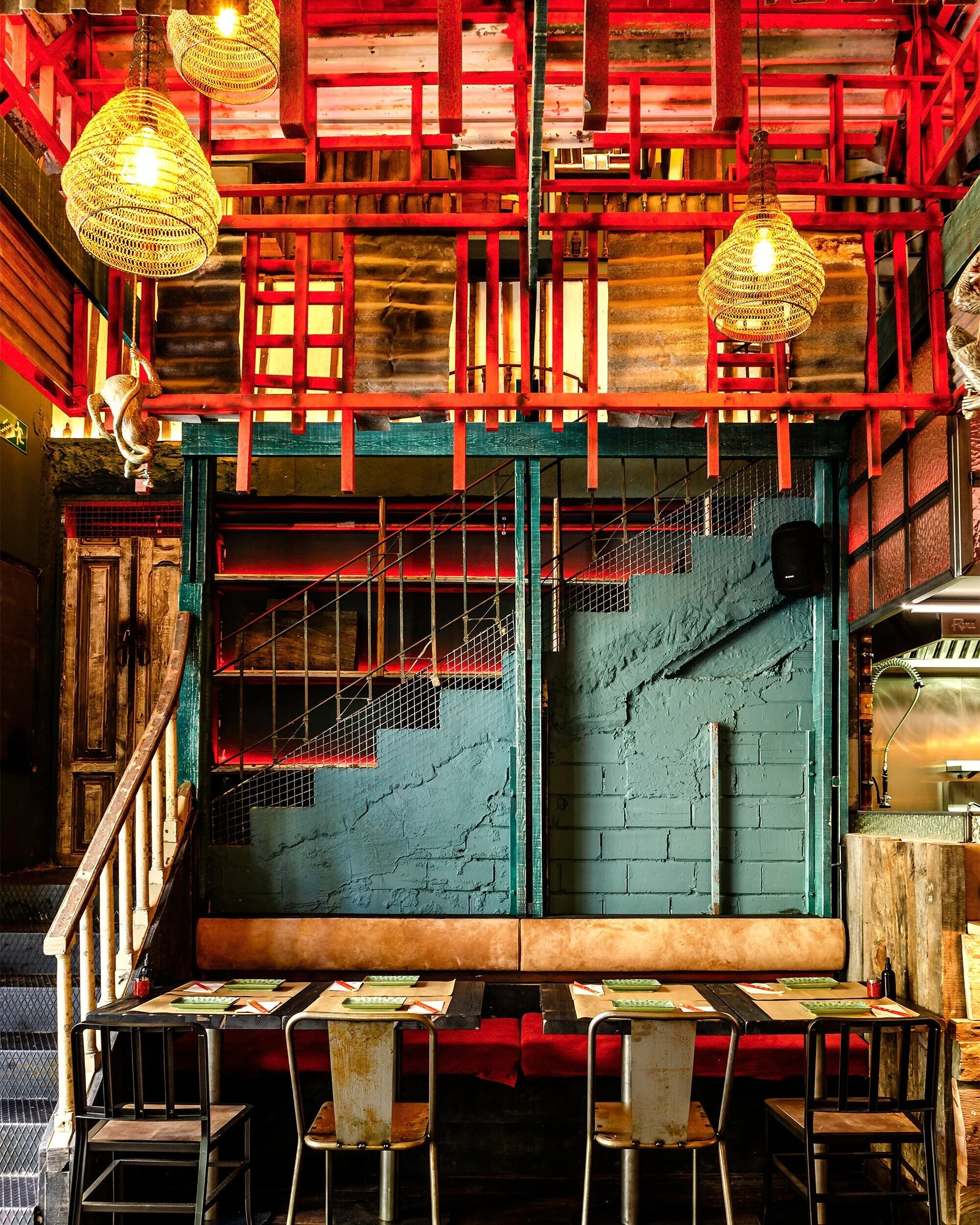 Interior of a restaurant with a green brick wall, a staircase with red railing, hanging wicker lamps, and a seating area with a table set for four.